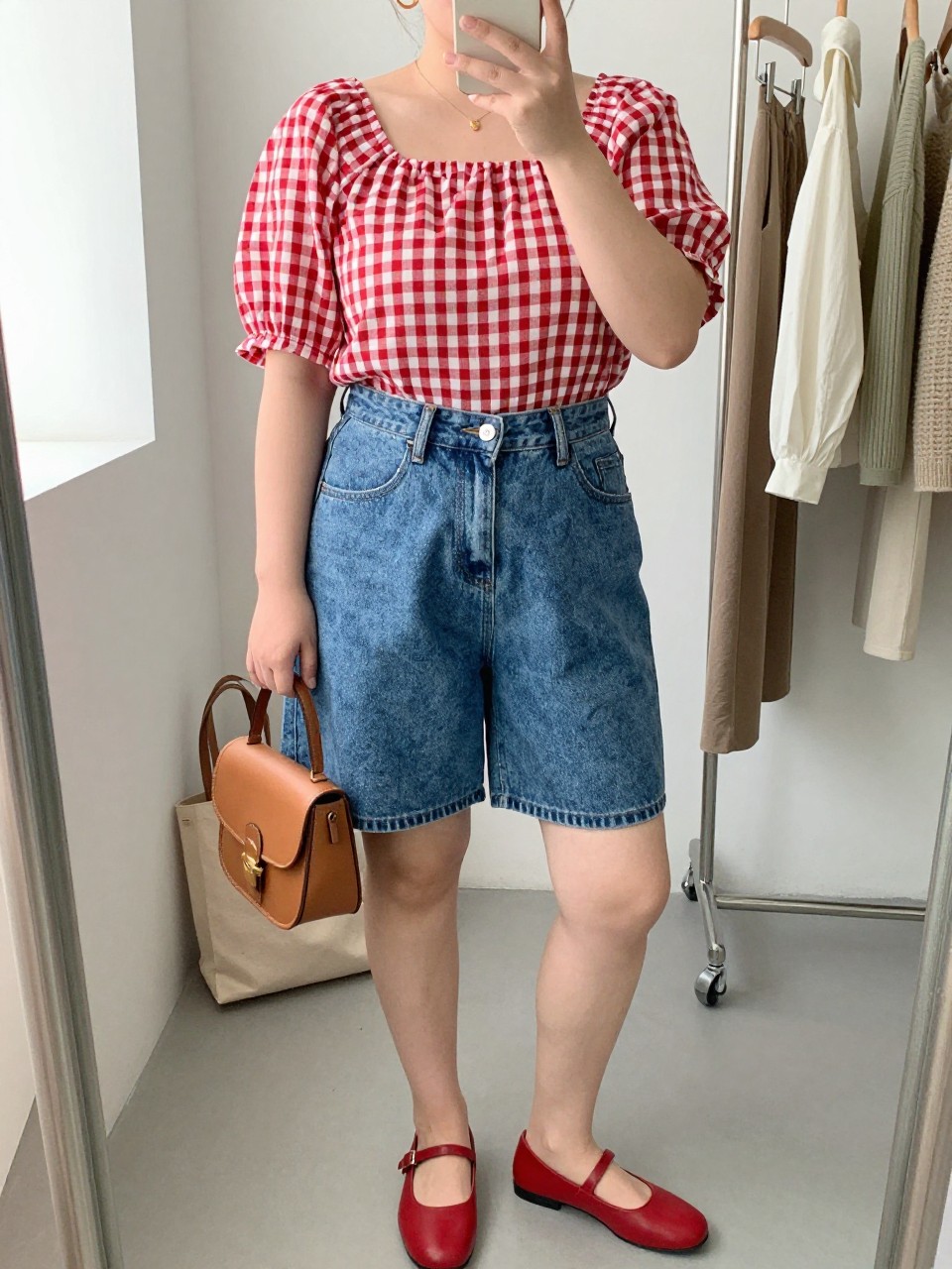 Mirror selfie of a plus size woman wearing medium-wash high-waisted denim shorts with a relaxed mid-thigh cut, a red and white gingham blouse with short puff sleeves and a square neckline, red flat Mary Jane shoes, a small tan leather saddle bag, and delicate gold hoop earrings, clearly showing the exact clothing items, denim wash, gingham print, sleeve shape, footwear, and accessories listed, holding a phone in front of a mirror, cropped at the neck so her face is hidden, relaxed playful selfie pose, in a fashion studio mirror corner with natural window light, a clothing rack with garments behind her and a tote bag resting on the floor, realistic picnic-ready summer outfit, latest iPhone photo quality.