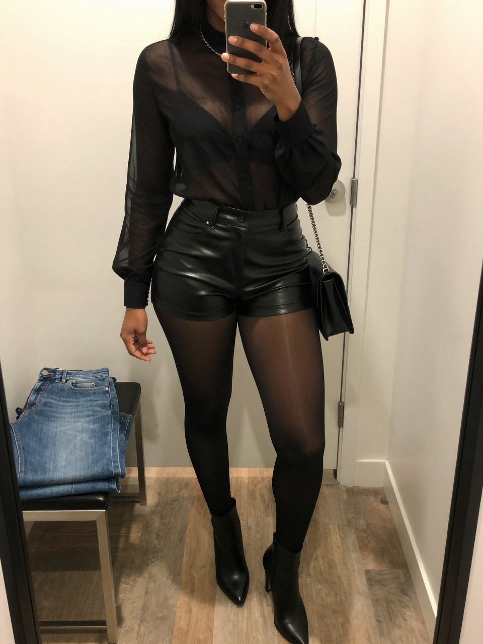 mirror selfie of a Black woman wearing the exact outfit described above, clearly showing a sheer black long-sleeve blouse over a black bralette, black faux leather high-waisted shorts, black sheer tights, black heeled ankle boots, and black chain shoulder bag, holding a phone in front of a mirror, cropped at NECK so the face does not show, with a confident casual selfie pose, in a walk-in closet mirror, soft indoor lighting, including folded denim jeans on a bench and a handbag hanging on a door handle, latest iPhone photo quality.