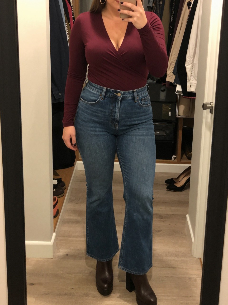 Mirror selfie of a plus size woman wearing high-rise medium-wash flared jeans, a burgundy long-sleeve wrap top, gold hoop earrings, and dark brown platform ankle boots, clearly showing the exact outfit pieces, colors, fabrics, and accessories listed, holding a phone in front of a mirror, cropped at the neck so the face does not show, relaxed selfie stance, in a walk-in closet mirror with warm moody lighting, an open closet with clothes visible and a pair of heels on the floor nearby, realistic casual composition, latest iPhone photo quality.