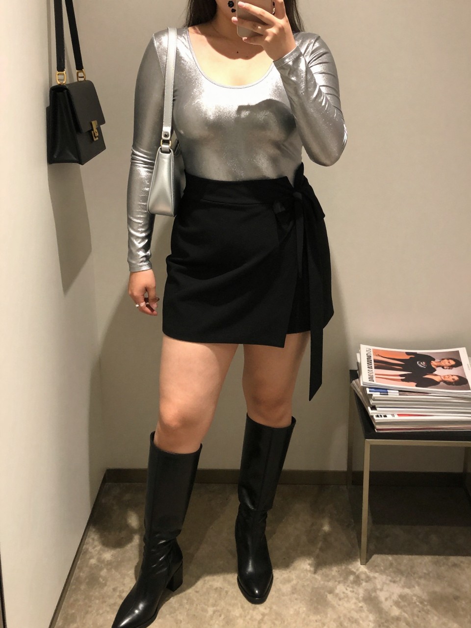 Mirror selfie of a plus size woman wearing a silver metallic long-sleeve bodysuit with a soft scoop neckline, a high-waisted black wrap mini skirt with an adjustable side tie, black knee-high boots with a low block heel, a compact silver shoulder bag, and black crystal stud earrings, clearly showing the exact clothing pieces, metallic finish, wrap detail, colors, footwear, and accessories listed, holding a phone in front of a mirror, cropped at the neck so the face is hidden, relaxed evening selfie pose, in a minimal dressing room with warm moody lighting, a handbag hanging on a door handle and a stack of fashion magazines on a side table, realistic nightclub styling, latest iPhone photo quality.