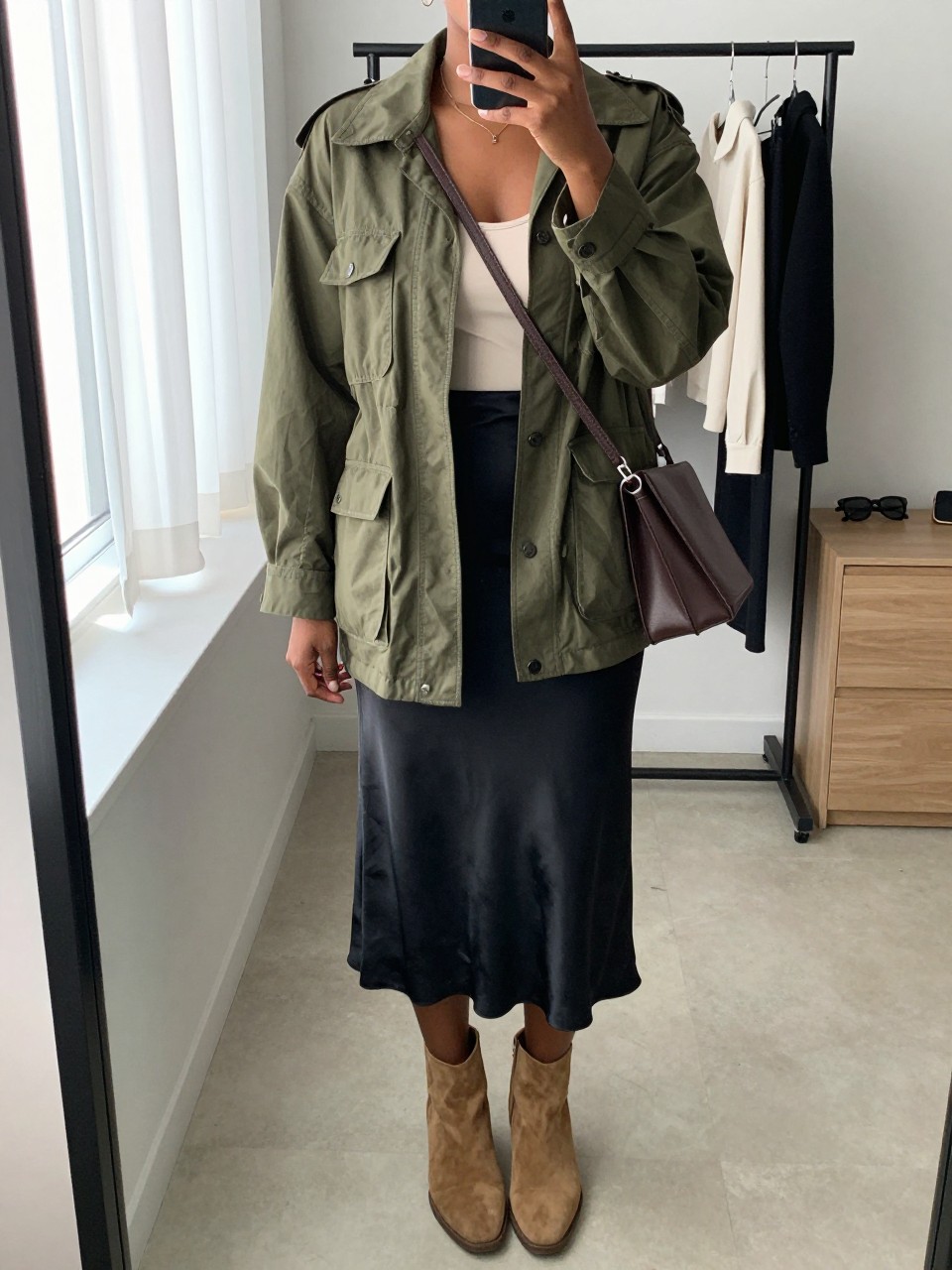Mirror selfie of a Black woman wearing an olive-green lightweight utility jacket over a fitted ivory camisole, black satin midi slip skirt, tan suede ankle boots, and a structured chocolate-brown crossbody bag, clearly showing the exact clothing items, colors, fabrics, and accessories listed, holding a phone in front of a minimal dressing room mirror, cropped at the neck so the face does not show, relaxed casual selfie pose, natural window light, a clothing rack with garments behind and sunglasses placed on a dresser, polished yet natural, latest iPhone photo quality.
