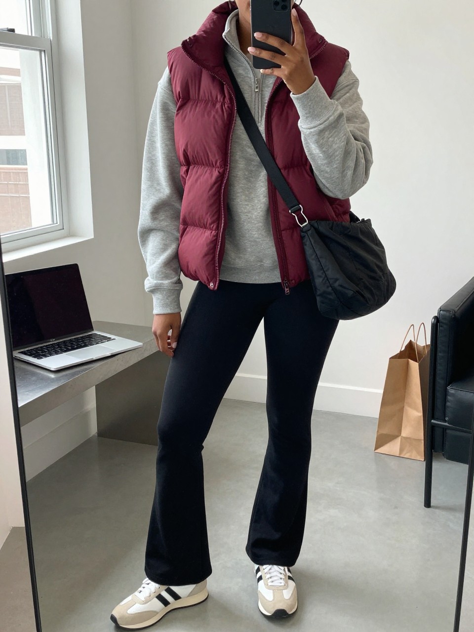 Mirror selfie of a Black woman wearing a burgundy quilted puffer vest over a fitted heather-grey quarter-zip sweatshirt, black flared leggings, white and beige retro sneakers, and a black nylon crossbody bag, clearly showing the exact clothing items, colors, fabrics, and accessories listed, holding a phone in front of a fashion studio mirror corner, cropped at the neck so the face does not show, relaxed casual selfie pose, natural window light, a laptop on a bench and shopping bags beside a chair, believable casual setting, latest iPhone photo quality.