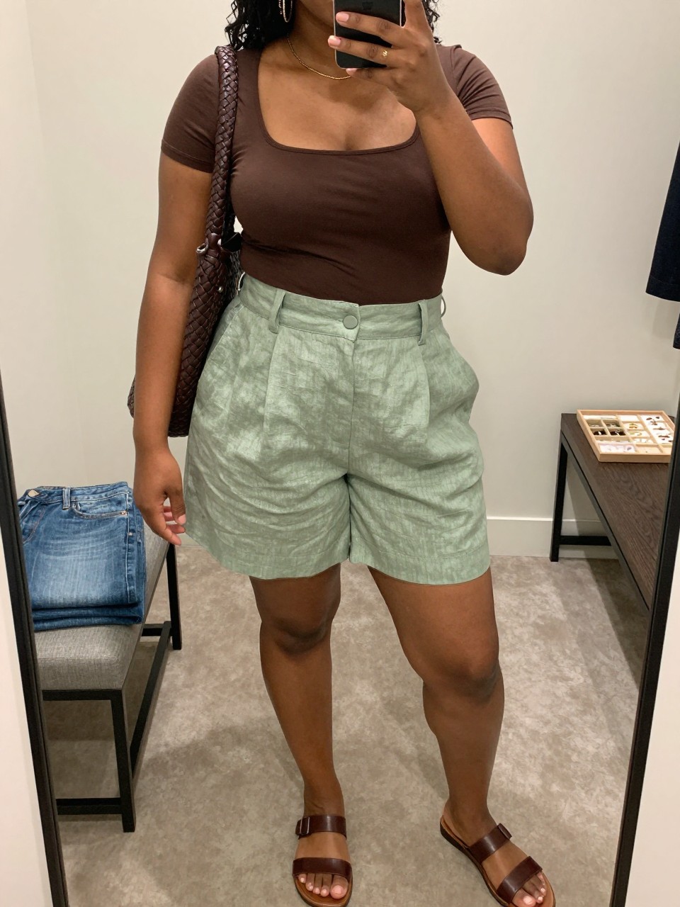 Mirror selfie of a black plus size woman wearing sage green high-waisted linen-blend shorts with a softly tailored shape, a fitted chocolate brown square-neck short-sleeve top, dark brown flat leather sandals, a chocolate woven shoulder bag, and delicate gold chain earrings, clearly showing the exact clothing items, sage fabric, chocolate top, colors, footwear, and accessories listed, holding a phone in front of a mirror, cropped at the neck so the face is hidden, relaxed polished selfie pose, in a walk-in closet mirror with soft indoor lighting, folded denim jeans on a bench and a jewelry tray on the vanity, realistic earthy summer outfit, latest iPhone photo quality.