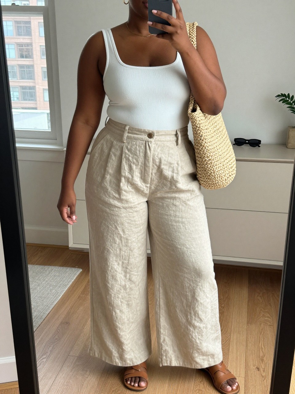 Mirror selfie of a black plus size woman wearing beige high-waisted wide-leg linen pants with soft front pleats, a fitted white ribbed square-neck bodysuit, tan flat leather sandals, a natural woven shoulder bag, and delicate gold hoop earrings, clearly showing the exact clothing items, beige linen, white ribbed fabric, colors, footwear, and accessories listed, holding a phone in front of a mirror, cropped at the neck so the face does not show, relaxed casual selfie pose, in a stylish city apartment with natural window light, sunglasses placed on a dresser and a small plant nearby, natural unstaged warm-weather appearance, latest iPhone photo quality.