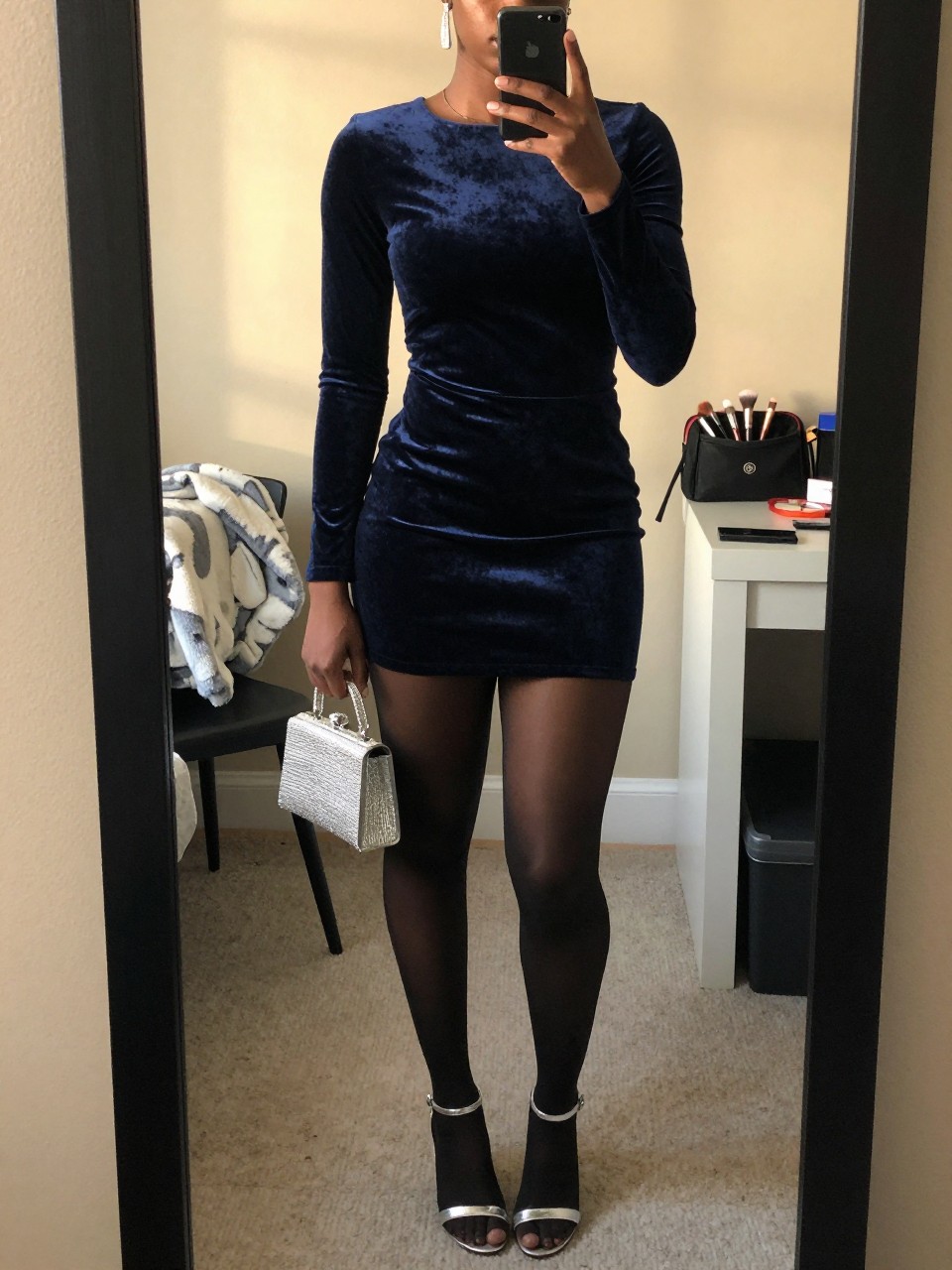 mirror selfie of a Black woman wearing the exact outfit described above, clearly showing a navy velvet long-sleeve mini dress, silver strappy heels, black sheer tights, silver crystal clutch, and silver drop earrings, holding a phone in front of a mirror, cropped at NECK so the face does not show, with a confident casual selfie pose, in a feminine-themed bedroom, evening golden hour, including a folded blanket on a chair and a makeup brush or cosmetic bag on the counter, latest iPhone photo quality.