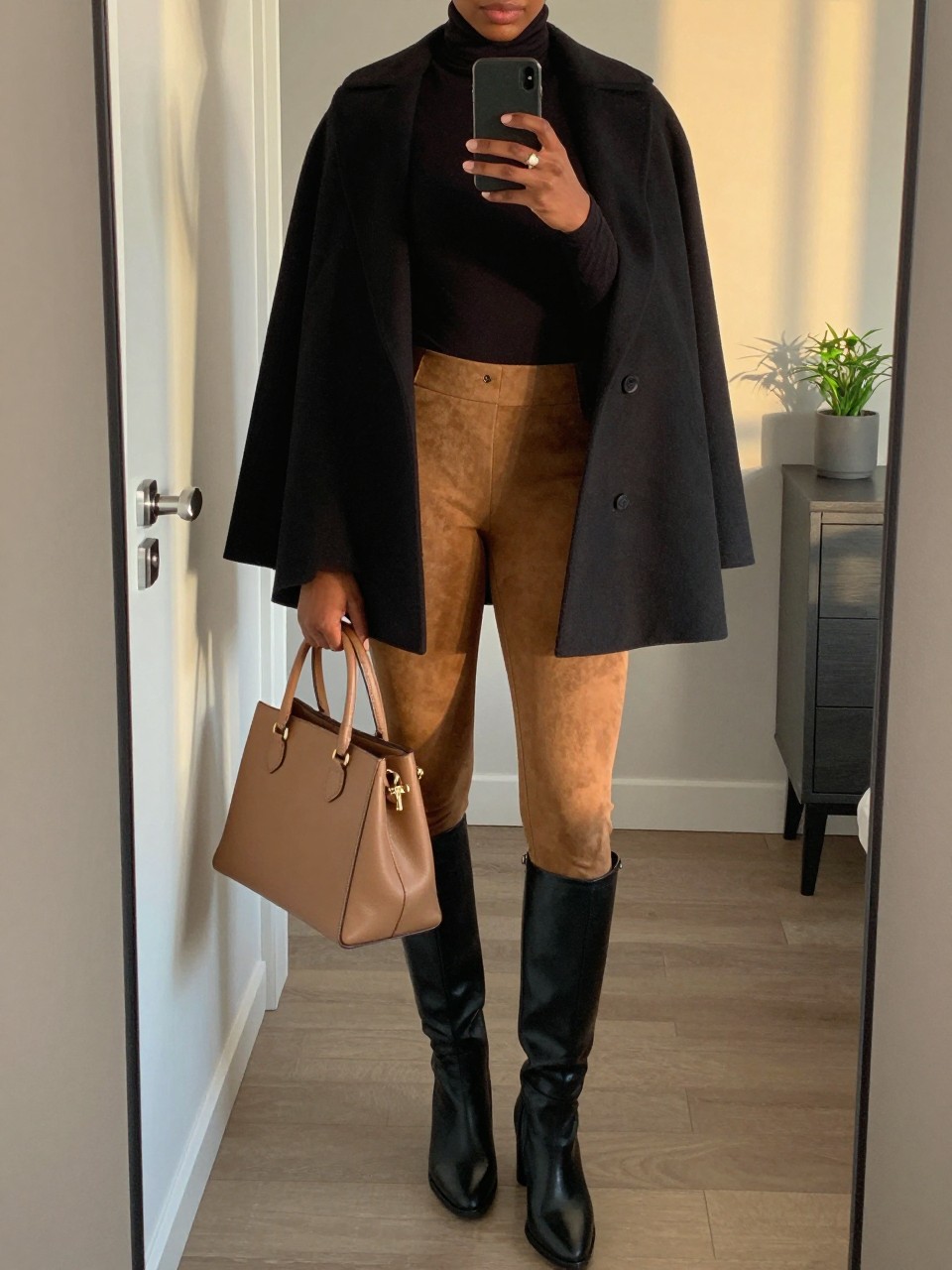 Mirror selfie of a Black woman wearing camel faux-suede leggings, a fitted black turtleneck, a structured black wool cape coat, black knee-high leather boots, and a camel top-handle handbag, clearly showing the exact clothing items, colors, fabrics, and accessories listed, holding a phone in front of a stylish city apartment mirror, cropped at the neck so the face does not show, relaxed casual selfie pose, evening golden-hour lighting, handbag hanging on a door handle and a small plant on a dresser, elegant natural composition, latest iPhone photo quality.