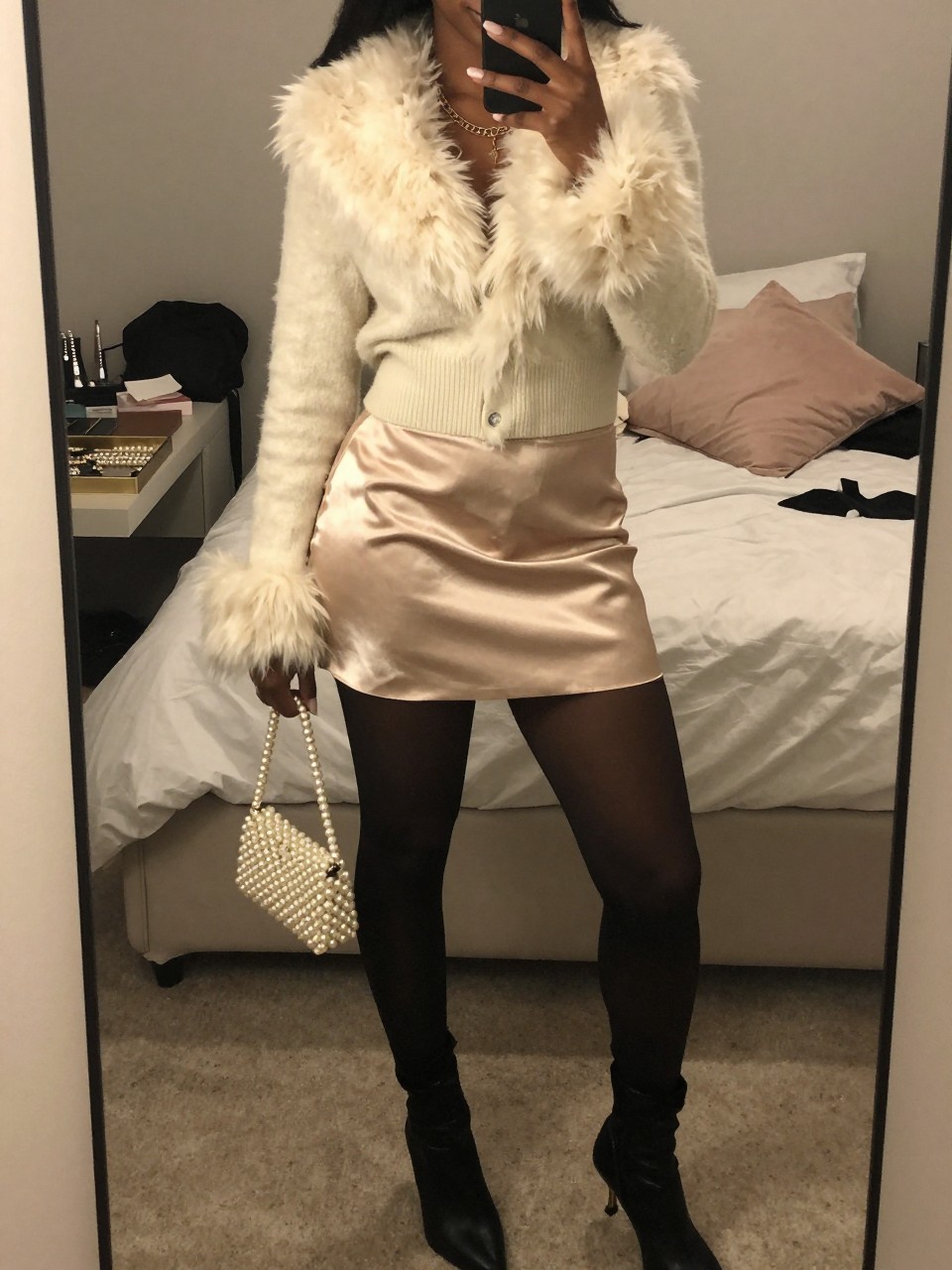 mirror selfie of a Black woman wearing a cream fur-trim cardigan, champagne satin mini skirt, sheer black tights, black kitten heel boots, and a pearl mini shoulder bag, clearly showing the same clothing items, colors, fabrics, and accessories listed in the Outfit Pieces section, holding a phone in front of a mirror, cropped at NECK so the face does not show, with a relaxed casual selfie pose, in a feminine-themed bedroom, warm moody lighting, including soft pillows on an unmade bed and a jewelry tray on the vanity, latest iPhone photo quality.