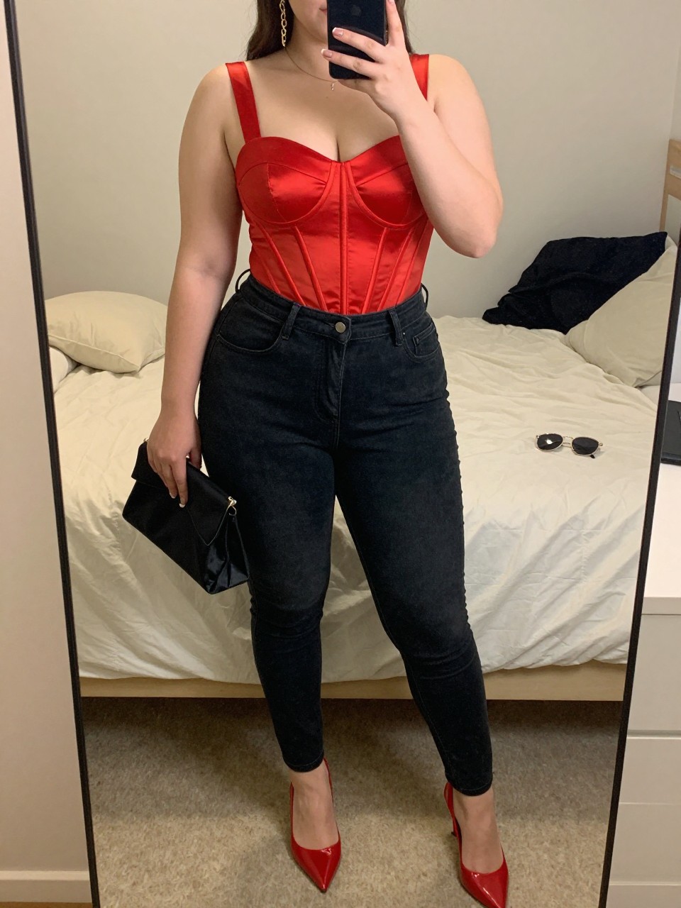 Mirror selfie of a plus size woman wearing a red satin corset-style top with a structured bodice and wide straps, high-waisted black coated skinny jeans, red pointed slingback heels, a small black satin clutch, and gold chain earrings, clearly showing the exact clothing items, red satin, coated denim, colors, footwear, and accessories listed, holding a phone in front of a mirror, cropped at the neck so the face does not appear, relaxed polished selfie stance, in a cozy bedroom mirror setup with soft indoor lighting, soft pillows on an unmade bed and sunglasses placed on a dresser, realistic club date-night outfit, latest iPhone photo quality.