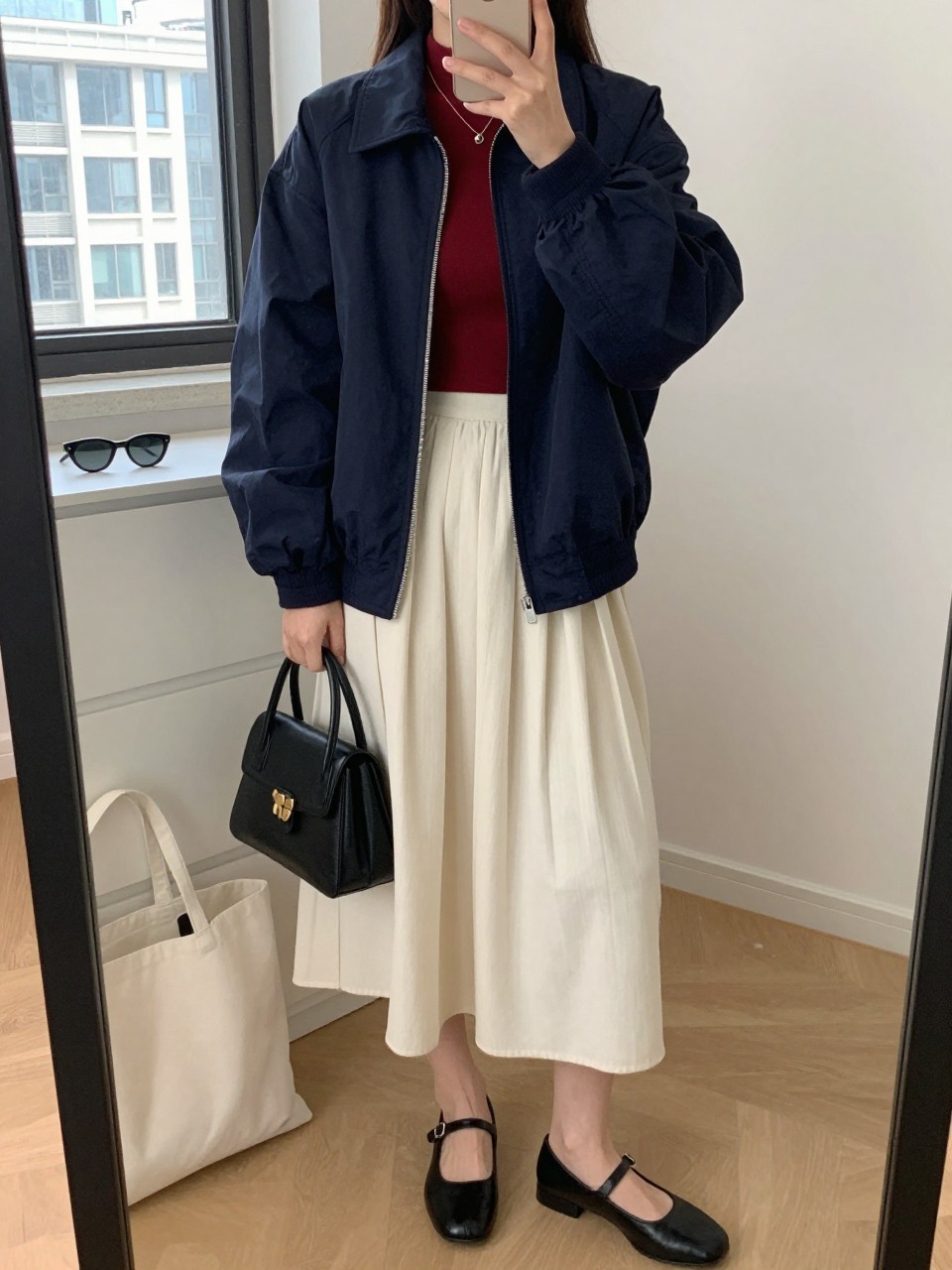 Mirror selfie of a woman wearing an oversized navy bomber jacket, fitted burgundy mock-neck top, cream pleated midi skirt, black leather Mary Jane shoes, and a small black top-handle bag, clearly showing the exact clothing items, colors, fabrics, and accessories listed, holding a phone in front of a stylish city apartment mirror, cropped at the neck so the face does not show, relaxed casual selfie pose, natural window light, sunglasses placed on a dresser and a tote bag resting on the floor, natural everyday setting, latest iPhone photo quality.