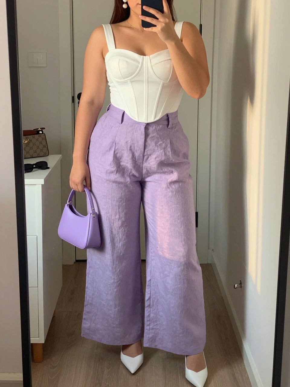Mirror selfie of a plus size woman wearing lavender high-waisted wide-leg linen trousers, a structured white corset-style top with wide supportive straps, white pointed slingback heels, a small lavender top-handle bag, and pearl drop earrings, clearly showing the exact clothing items, lavender linen, white structured top, colors, footwear, and accessories listed, holding a phone in front of a mirror, cropped at the neck so her face does not appear, relaxed feminine selfie pose, in a walk-in closet mirror with evening golden-hour lighting, sunglasses placed on a dresser and a handbag hanging on a door handle, realistic birthday brunch outfit, latest iPhone photo quality.