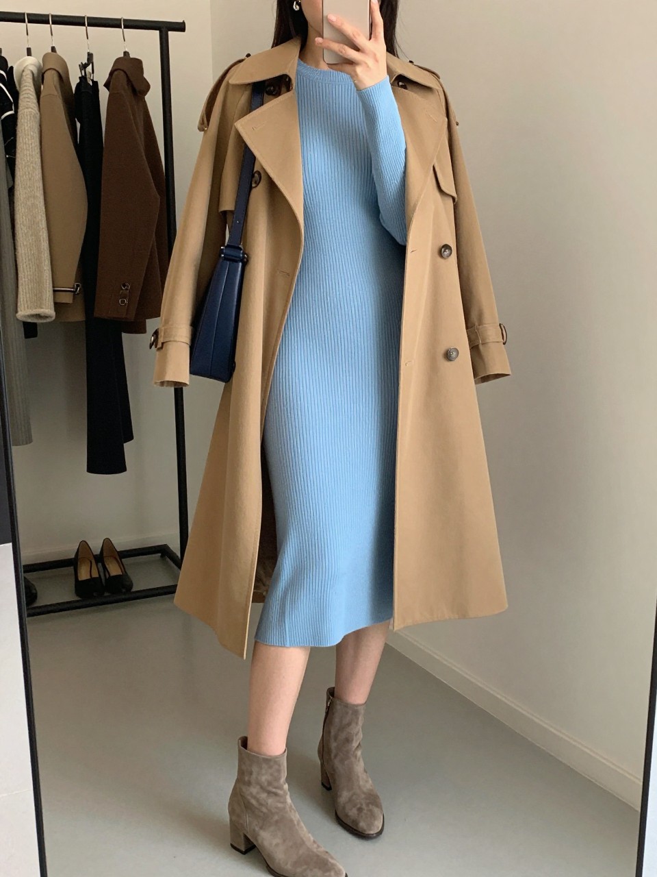 Mirror selfie of a woman wearing a fitted powder-blue ribbed midi sweater dress, knee-length camel trench coat worn open, taupe suede ankle boots, a structured navy shoulder bag, and small silver drop earrings, clearly showing the exact clothing items, colors, fabrics, and accessories listed, holding a phone in front of a fashion studio mirror corner, cropped at the neck so the face does not show, relaxed casual selfie pose, evening golden-hour lighting, a clothing rack with garments behind and a pair of heels on the floor nearby, elegant but natural, latest iPhone photo quality.