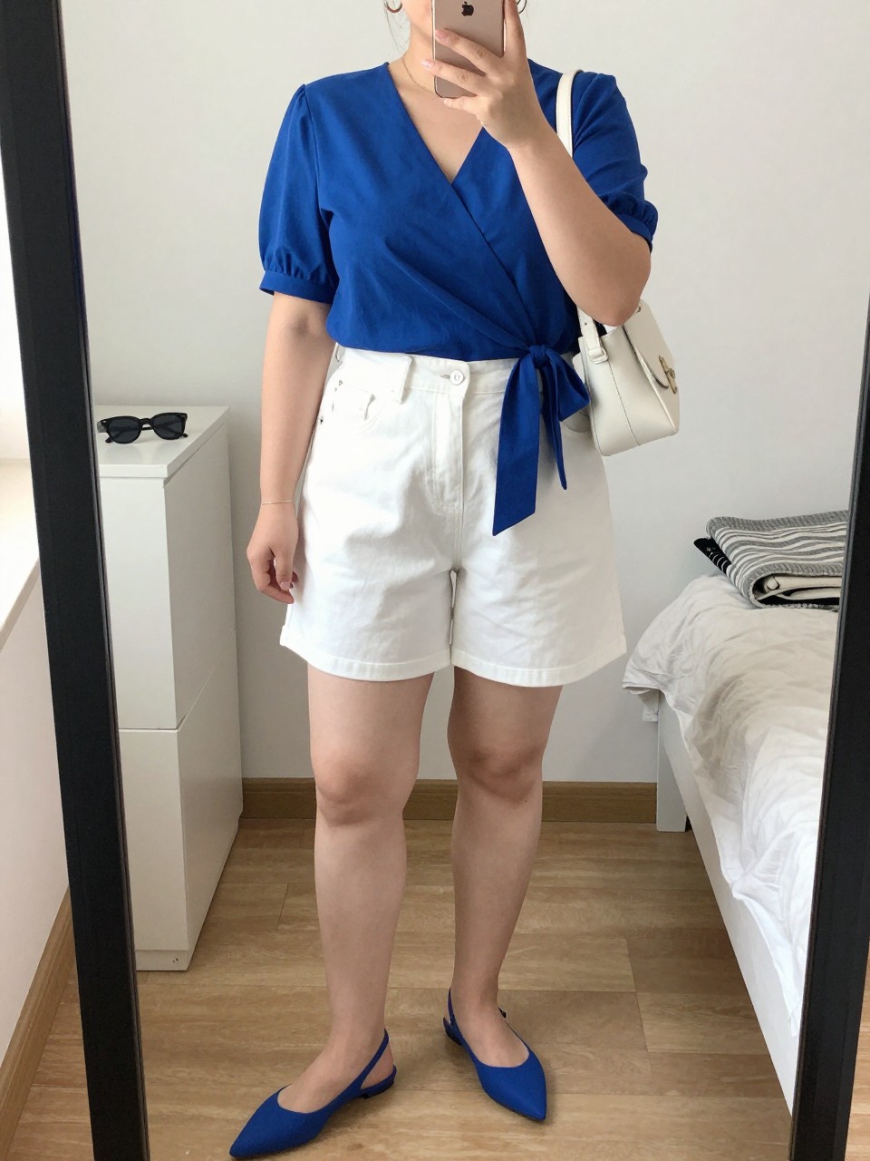 Mirror selfie of a plus size woman wearing crisp white high-waisted denim shorts with a clean mid-thigh hem, a cobalt blue short-sleeve wrap top with a softly tied waist, cobalt pointed flat slingbacks, a small white leather shoulder bag, and silver hoop earrings, clearly showing the exact clothing items, white denim, cobalt fabric, colors, footwear, and accessories listed, holding a phone in front of a mirror, cropped at the neck so the face does not appear, relaxed bright selfie stance, in a cozy bedroom mirror setup with natural window light, sunglasses placed on a dresser and a folded blanket on a chair, realistic summer lunch outfit, latest iPhone photo quality.