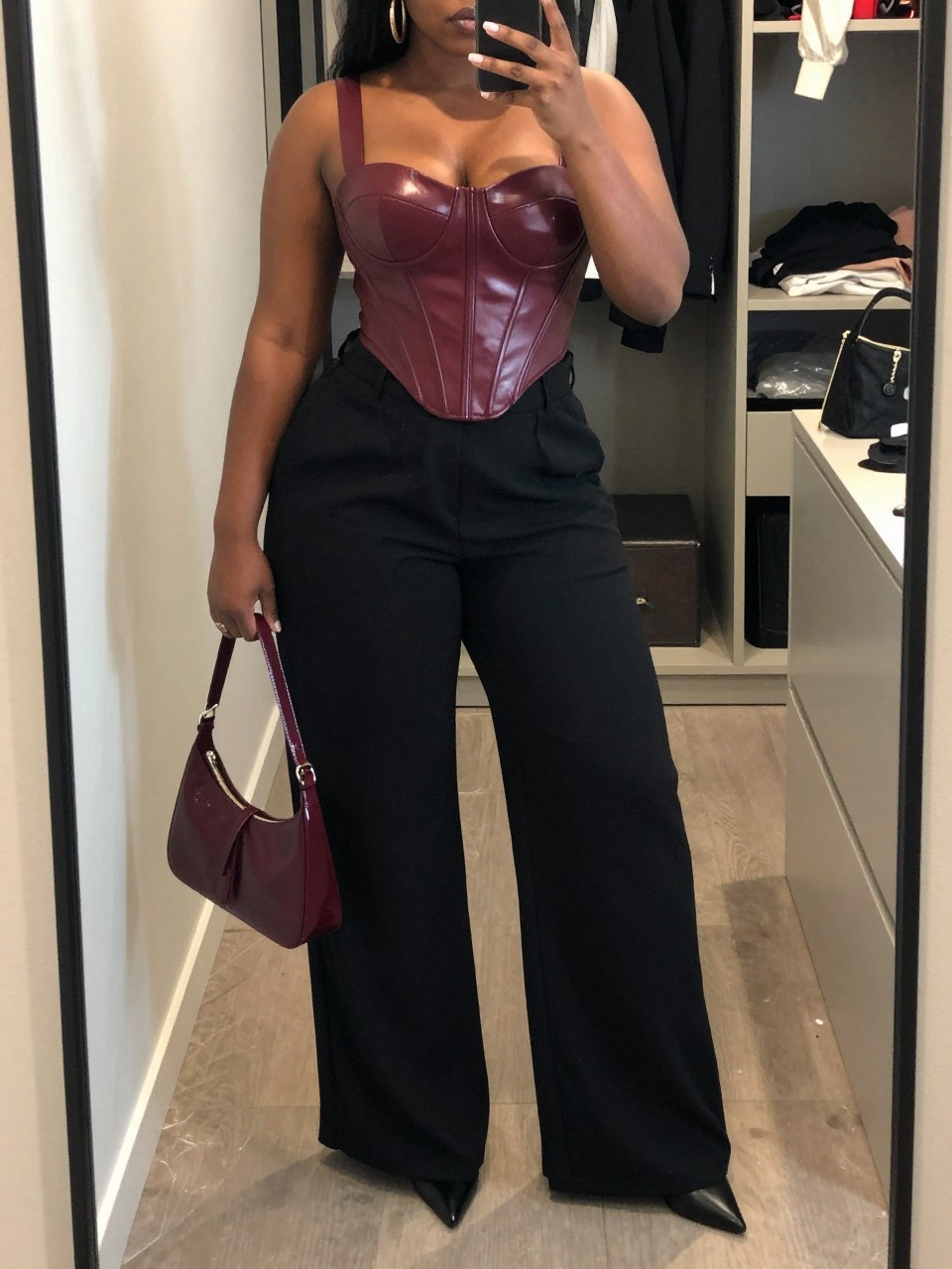 Mirror selfie of a black plus size woman wearing a structured burgundy faux-leather corset top with wide supportive straps, high-waisted black wide-leg trousers in fluid fabric, black pointed platform heels, a burgundy patent shoulder bag, and chunky gold hoop earrings, clearly showing the exact clothing items, colors, fabrics, faux-leather texture, footwear, and accessories listed, holding a phone in front of a mirror, cropped at the neck so her face does not appear, relaxed confident selfie stance, in a walk-in closet mirror with soft indoor lighting, an open closet with clothes visible and a cosmetic bag on the counter, realistic upscale club outfit, latest iPhone photo quality.