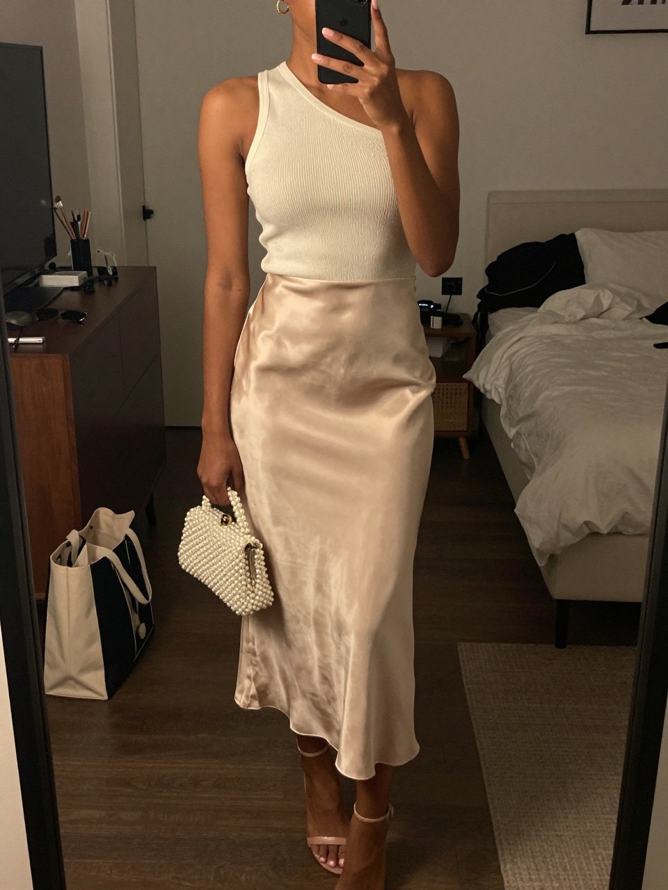 Image prompt mirror selfie of a black woman wearing a champagne satin slip midi skirt with an ivory one-shoulder knit top, nude strappy heels, a pearl-finish clutch bag, and small gold hoop earrings, holding a phone in front of a mirror, cropped at NECK so face doesn't show, with a natural casual selfie pose, in a city apartment, warm moody lighting, including small lived-in details like a tote bag on the floor, sunglasses on a dresser, or soft bedding in the background, latest iPhone photo quality.