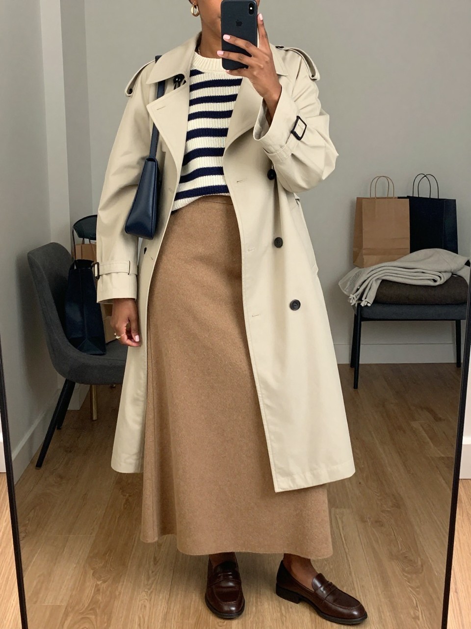 mirror selfie of a Black woman wearing the exact outfit described above, clearly showing a camel wool maxi skirt, navy and cream striped sweater, brown leather loafers, navy shoulder bag, gold huggie earrings, and a cream trench coat worn open, holding a phone in front of a mirror, cropped at NECK so the face does not show, with a relaxed casual selfie pose, in a stylish city apartment, soft indoor lighting, with shopping bags beside a chair and folded blanket on a chair, latest iPhone photo quality.