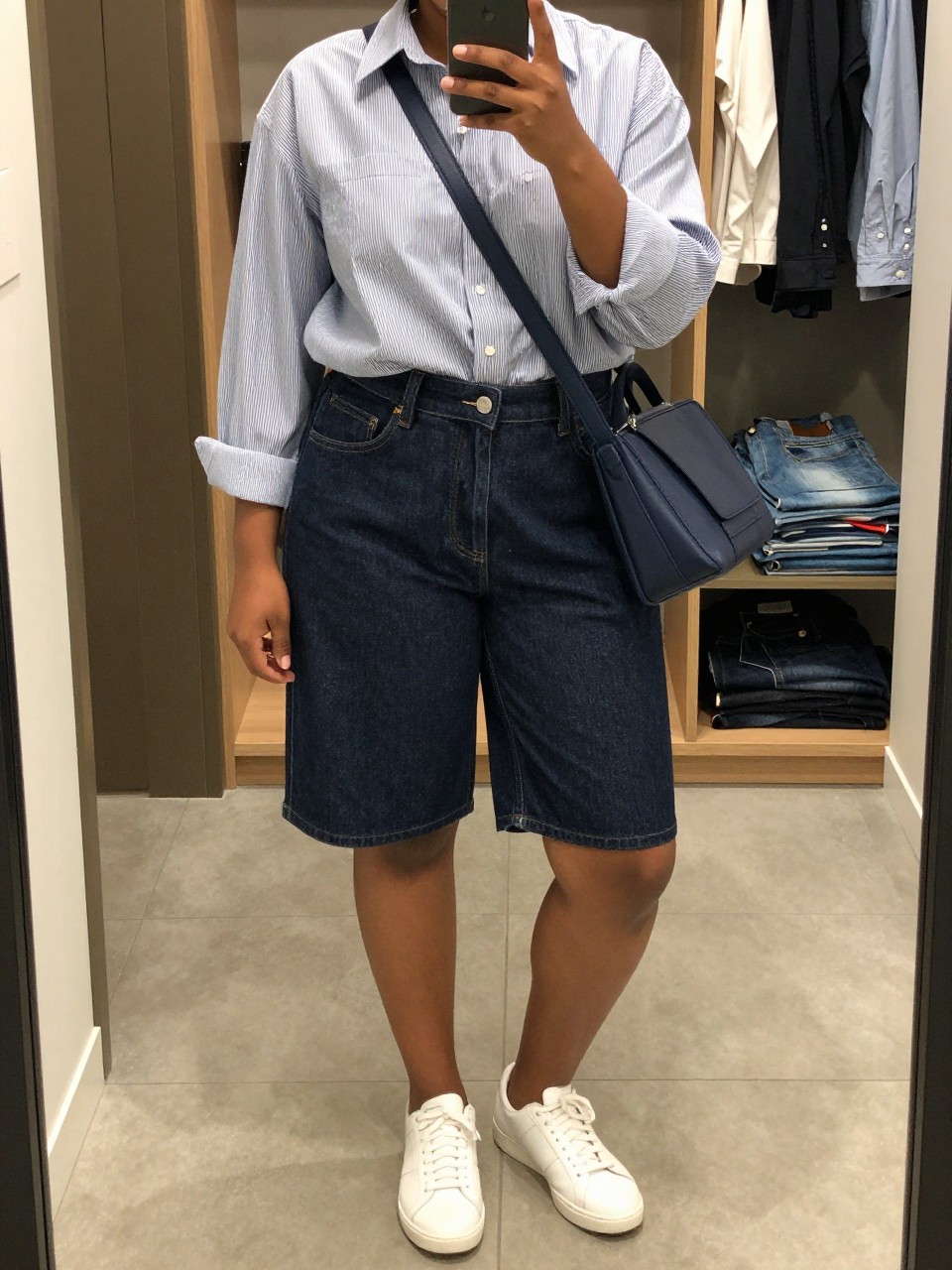 Mirror selfie of a black plus size woman wearing dark indigo high-waisted denim Bermuda shorts ending just above the knees, an oversized blue and white striped cotton button-down shirt tucked loosely at the front, white leather sneakers, and a structured navy crossbody bag, clearly showing the exact clothing items, denim wash, stripe pattern, colors, fabrics, footwear, and accessories listed, holding a phone in front of a mirror, cropped at the neck so her face does not appear, relaxed polished selfie stance, in a walk-in closet mirror with soft indoor lighting, an open closet with clothes visible and folded denim jeans on a bench, realistic everyday summer styling, latest iPhone photo quality.