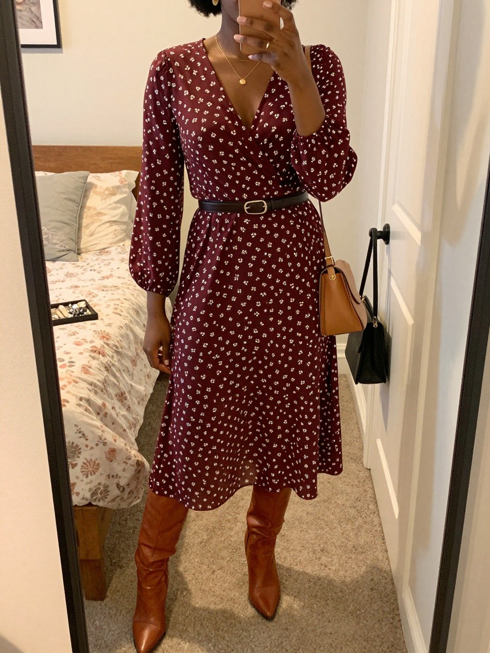 Mirror selfie of a Black woman wearing a burgundy long-sleeve wrap midi dress with a small cream floral print, cognac leather knee-high boots, a slim dark brown leather belt, tan saddle bag, and small hammered-gold earrings, clearly showing the exact clothing items, colors, fabrics, and accessories listed, holding a phone in front of a feminine-themed bedroom mirror, cropped at the neck so the face does not show, relaxed casual selfie pose, evening golden-hour lighting, a jewelry tray on the vanity and a handbag hanging on a door handle, warm and natural, latest iPhone photo quality.