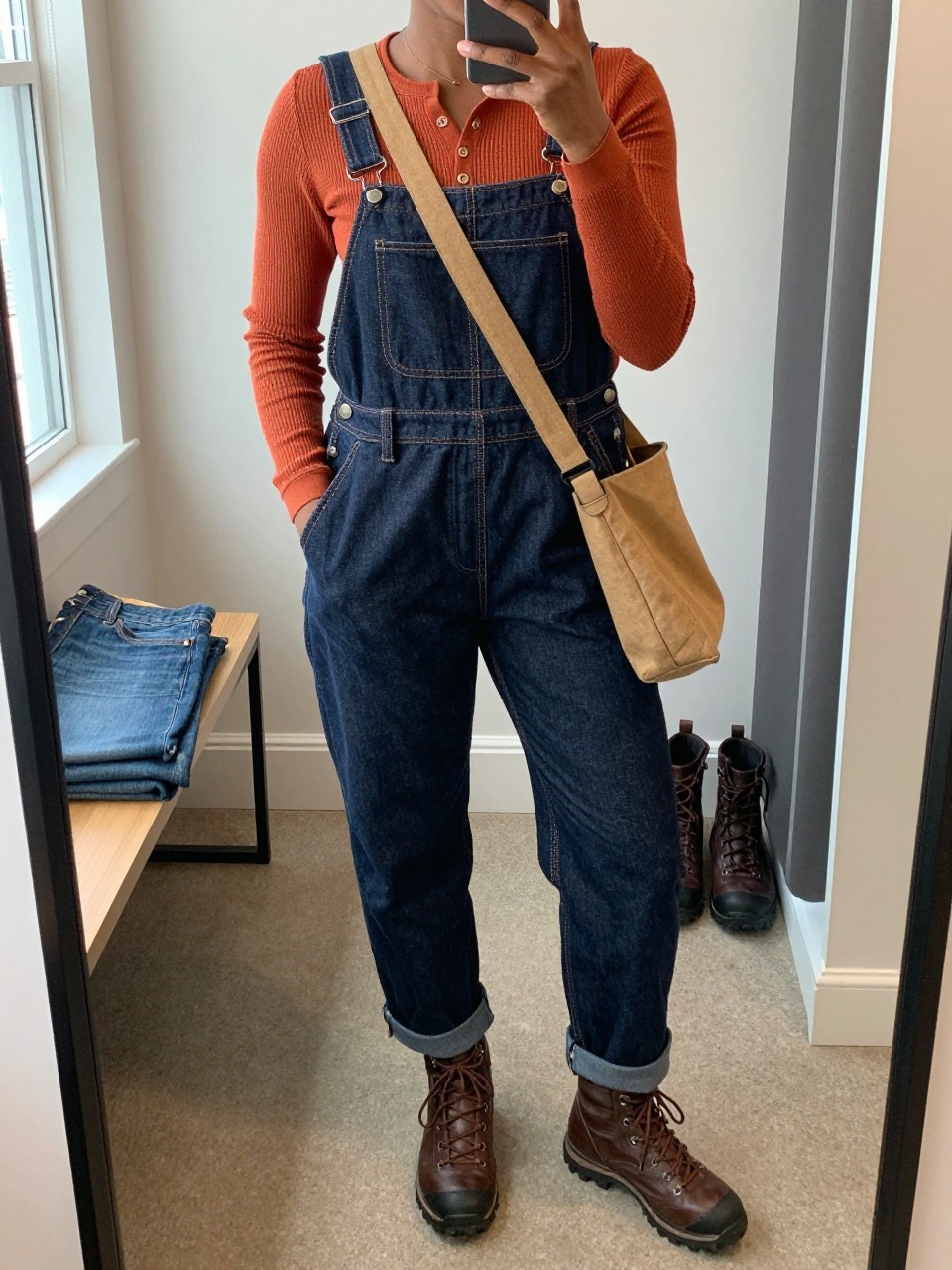 Mirror selfie of a Black woman wearing dark indigo relaxed-fit denim overalls over a rust-orange waffle-knit thermal henley, chocolate-brown lace-up hiking boots, and a tan canvas crossbody bag, clearly showing the exact clothing items, colors, fabrics, and accessories listed, holding a phone in front of a walk-in closet mirror, cropped at the neck so the face does not show, relaxed casual selfie pose, natural window light, folded denim jeans on a bench and a pair of boots on the floor nearby, believable everyday setting, latest iPhone photo quality.