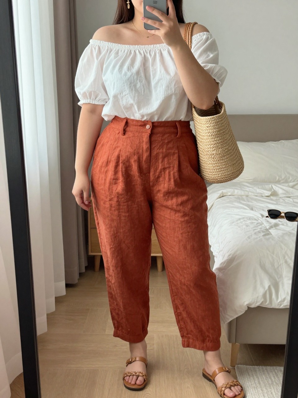 Mirror selfie of a plus size woman wearing terracotta high-waisted linen trousers with relaxed tapered legs, a white cotton off-shoulder blouse with short gathered sleeves, tan flat woven sandals, a natural straw shoulder bag, and delicate gold drop earrings, clearly showing the exact clothing items, terracotta linen, white cotton, gathered neckline, footwear, and accessories listed, holding a phone in front of a mirror, cropped at the neck so her face does not show, relaxed feminine selfie pose, in a cozy bedroom mirror setup with natural window light, soft pillows on an unmade bed and sunglasses placed on a dresser, realistic holiday outfit, latest iPhone photo quality.