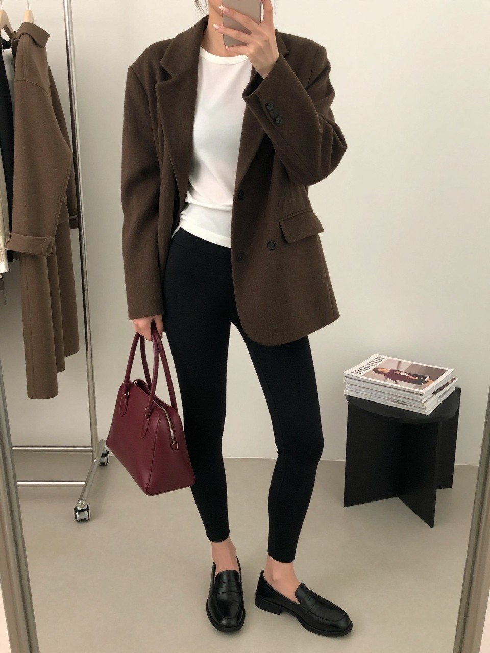 Mirror selfie of a woman wearing black stirrup leggings, a fitted white crew-neck top, an oversized chocolate-brown wool blazer, black leather loafers, and a burgundy top-handle bag, clearly showing the exact clothing items, colors, fabrics, and accessories listed, holding a phone in front of a minimal dressing room mirror, cropped at the neck so the face does not show, relaxed casual selfie pose, soft indoor lighting, clothing rack with garments behind and a stack of fashion magazines on a side table, natural editorial setting, latest iPhone photo quality.