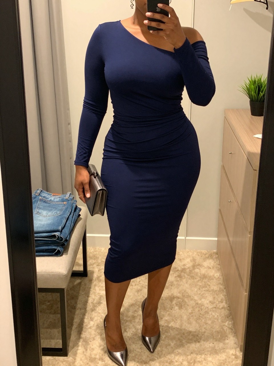 Mirror selfie of a black plus size woman wearing a deep midnight blue fitted midi dress with one long sleeve, an asymmetric neckline, and gentle ruching across the waist, metallic gunmetal pointed pumps, a small gunmetal box clutch, and silver spiral earrings, clearly showing the exact dress shape, blue color, ruching, footwear, and accessories listed, holding a phone in front of a mirror, cropped at the neck so her face does not appear, relaxed elegant selfie pose, in a walk-in closet mirror with evening golden-hour lighting, folded denim jeans on a bench and a small plant on a dresser, realistic sophisticated nightclub outfit, latest iPhone photo quality.