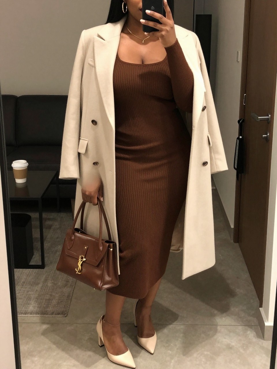 Mirror selfie of a black plus size woman wearing a chocolate brown ribbed knit midi dress with long sleeves, a square neckline, and a softly fitted shape, a cream longline blazer worn open, cream pointed block heels, a structured chocolate handbag, and hammered gold hoop earrings, clearly showing the exact clothing items, chocolate knit, cream blazer, footwear, and accessories listed, holding a phone in front of a mirror, cropped at the neck so the face does not appear, relaxed sophisticated selfie pose, in a stylish city apartment with warm moody lighting, a coffee mug on a side table and a handbag hanging on a door handle, realistic upscale dinner-date outfit, latest iPhone photo quality.