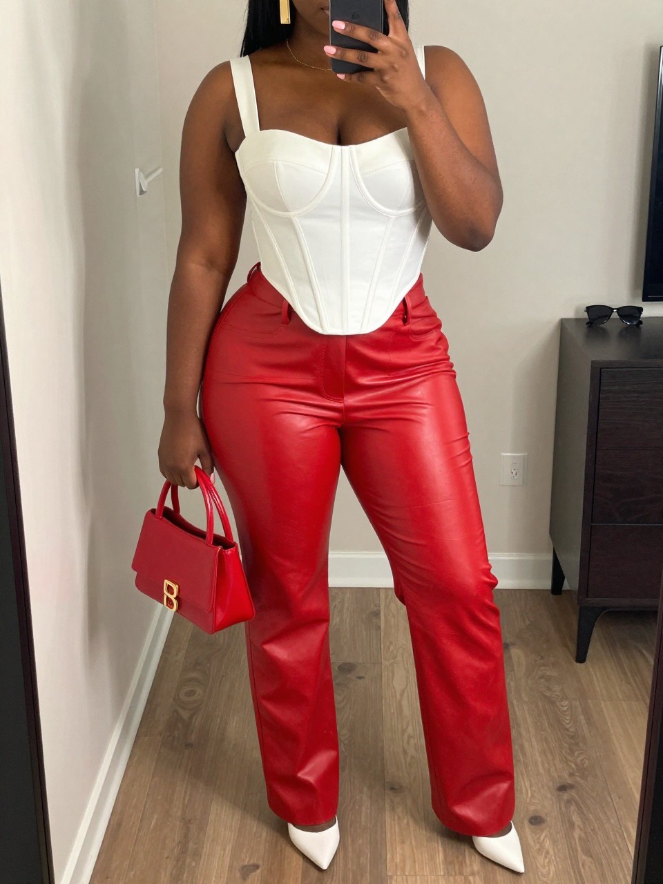 Mirror selfie of a black plus size woman wearing a structured white corset top with wide straps and a curved hem, high-waisted red faux-leather straight-leg pants, white pointed pumps, a small red patent handbag, and gold rectangular earrings, clearly showing the exact clothing items, white corset, red faux leather, colors, footwear, and accessories listed, holding a phone in front of a mirror, cropped at the neck so the face does not appear, relaxed confident selfie stance, in a stylish city apartment with soft indoor lighting, a handbag hanging on a door handle and sunglasses placed on a dresser, realistic bold club outfit, latest iPhone photo quality.