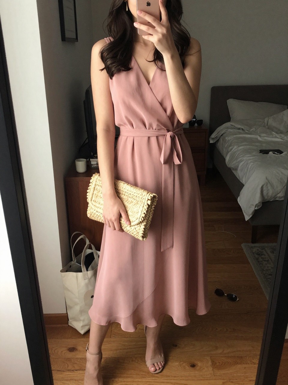 Image prompt mirror selfie of a woman wearing a dusty rose chiffon wrap midi dress with nude heeled sandals, a natural straw raffia clutch, simple gold drop earrings, and loose waves with a soft side part, holding a phone in front of a mirror, cropped at NECK so face doesn't show, with a natural casual selfie pose, in a city apartment, warm moody lighting, including small lived-in details like a tote bag on the floor, sunglasses on a dresser, or soft bedding in the background, latest iPhone photo quality.