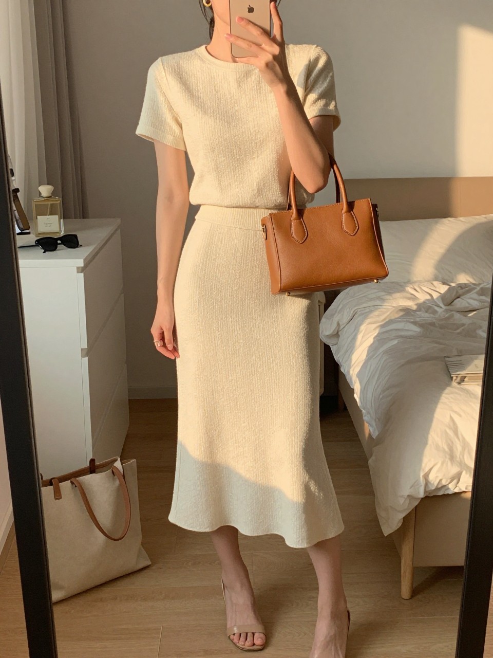 mirror selfie of a woman wearing a cream textured matching midi skirt set with nude heeled sandals, a tan leather structured mini bag, small gold hoop earrings, and hair half-up, holding a phone in front of a mirror, cropped at NECK so face doesn't show, with a natural casual selfie pose, in a feminine-themed bedroom, evening golden hour, including small lived-in details like a tote bag on the floor, sunglasses on a dresser, or soft bedding in the background, latest iPhone photo quality.