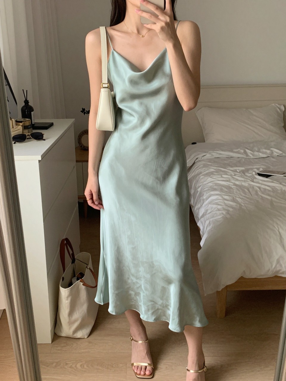 Image prompt mirror selfie of a woman wearing a soft seafoam green silk midi dress with champagne gold heeled sandals, a cream mini shoulder bag, a delicate gold necklace, and soft bronzy glowing makeup, holding a phone in front of a mirror, cropped at NECK so face doesn't show, with a natural casual selfie pose, in a feminine-themed bedroom, warm moody lighting, including small lived-in details like a tote bag on the floor, sunglasses on a dresser, or soft bedding in the background, latest iPhone photo quality.