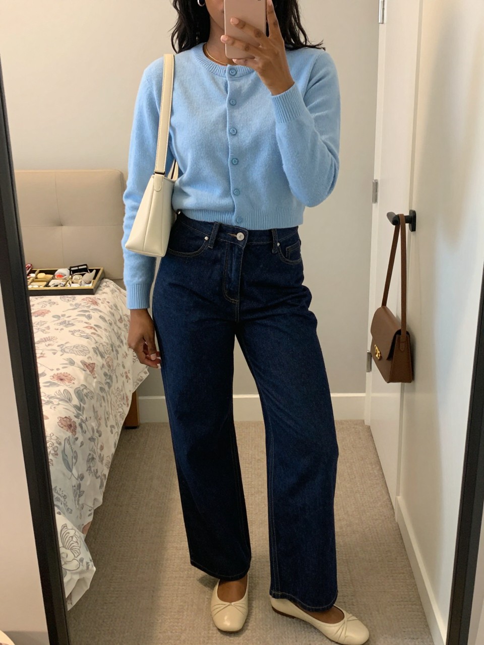 Mirror selfie of a Black woman wearing a cropped powder-blue button-front cardigan, high-waisted dark indigo wide-leg jeans, cream leather ballet flats, a structured ivory shoulder bag, and small silver hoop earrings, clearly showing the exact clothing items, colors, fabrics, and accessories listed, holding a phone in front of a feminine-themed bedroom mirror, cropped at the neck so the face does not show, relaxed casual selfie pose, soft indoor lighting, a jewelry tray on the vanity and a handbag hanging on a door handle, realistic and uncluttered, latest iPhone photo quality.