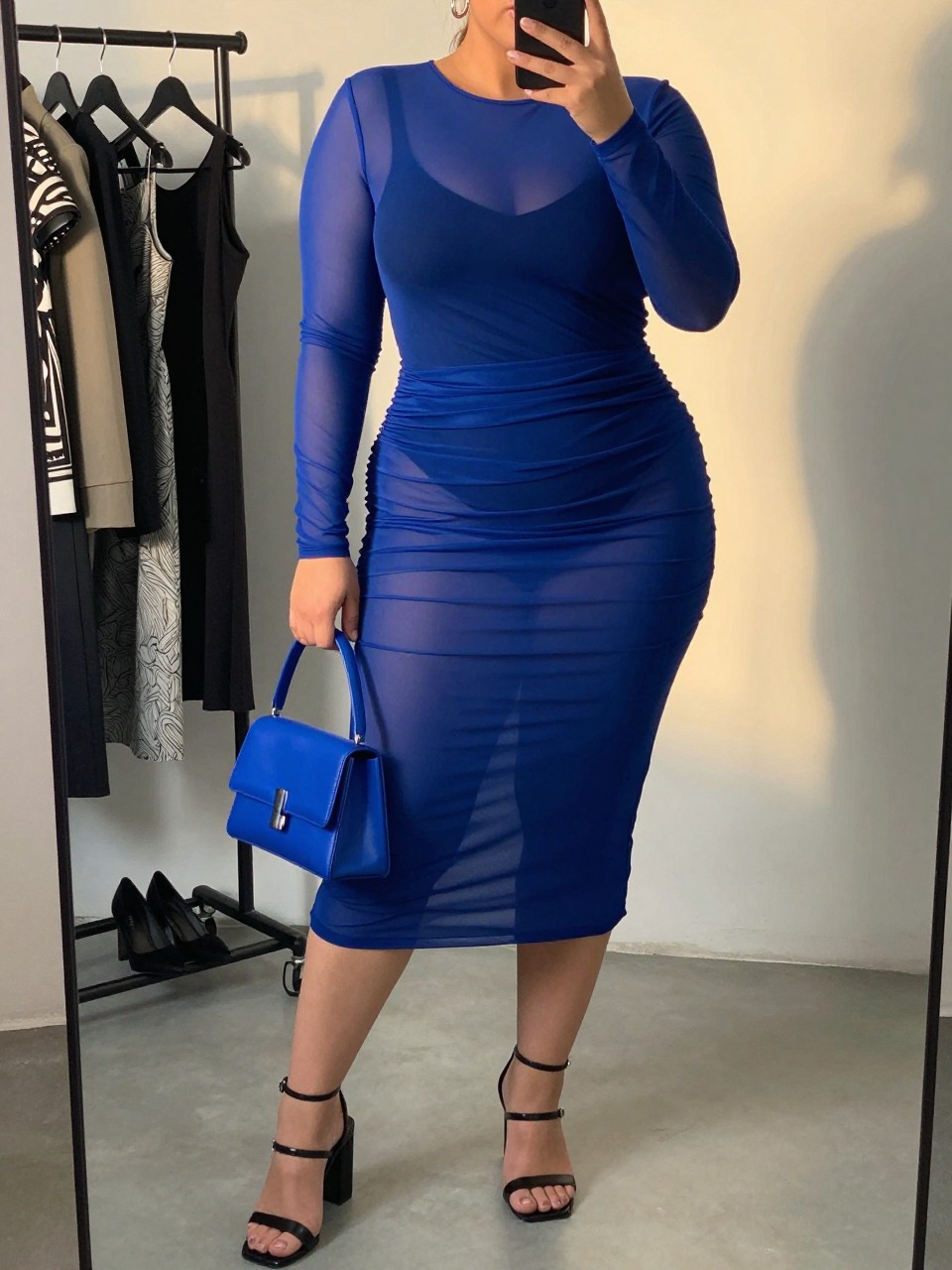 Mirror selfie of a plus size woman wearing a semi-sheer cobalt blue mesh midi dress with long sleeves and soft side gathering, layered over a fitted opaque black sleeveless bodysuit, black strappy block-heel sandals, a cobalt blue mini handbag, and silver hoop earrings, clearly showing the exact clothing layers, colors, mesh texture, gathering, footwear, and accessories listed, holding a phone in front of a mirror, cropped at the neck so her face does not show, relaxed confident selfie pose, in a fashion studio mirror corner with evening golden-hour lighting, a clothing rack with garments behind her and a pair of heels on the floor nearby, realistic bold club appearance, latest iPhone photo quality.