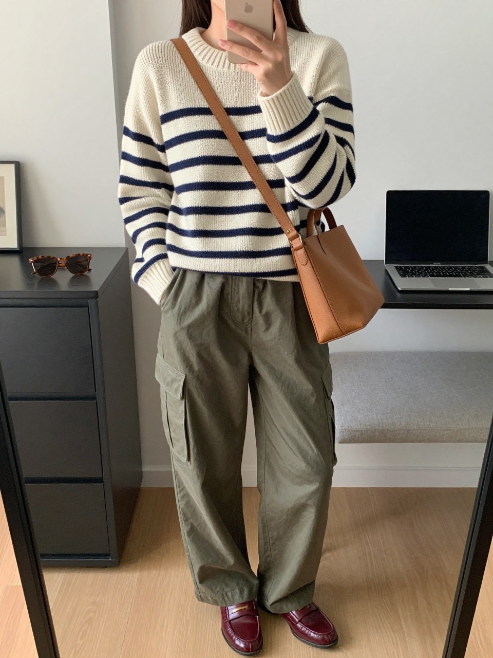 Mirror selfie of a woman wearing a cream and navy striped knit sweater, loose olive-green utility pants, burgundy leather loafers, a structured tan crossbody bag, and brown tortoiseshell sunglasses, clearly showing the exact clothing items, colors, fabrics, and accessories listed, holding a phone in front of a stylish city apartment mirror, cropped at the neck so the face does not show, relaxed casual selfie pose, soft indoor lighting, sunglasses placed on a dresser and a laptop on a bench, believable everyday setting, latest iPhone photo quality.
