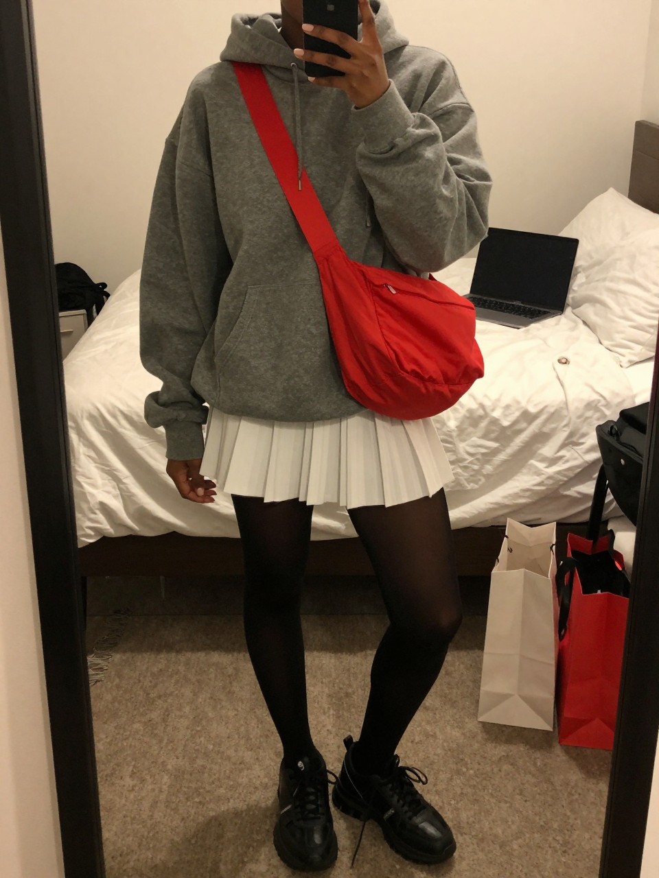 Mirror selfie of a Black woman wearing an oversized charcoal-grey hoodie, white pleated tennis mini skirt, opaque black tights, chunky black leather sneakers, and a vivid red nylon crossbody bag, clearly showing the exact clothing items, colors, fabrics, and accessories listed, holding a phone in front of a cozy bedroom mirror setup, cropped at the neck so the face does not show, relaxed casual selfie pose, warm moody lighting, a laptop on the bed and shopping bags beside a chair, casual lived-in atmosphere, latest iPhone photo quality.