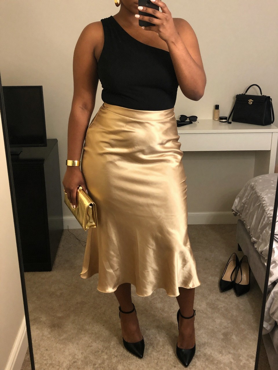 Mirror selfie of a black plus size woman wearing a metallic gold high-waisted satin midi skirt with a softly flared shape, a fitted black one-shoulder top with a secure wide strap, black pointed platform heels, a small gold hard-case clutch, and chunky gold cuff earrings, clearly showing the exact clothing items, gold satin, black asymmetric top, footwear, and accessories listed, holding a phone in front of a mirror, cropped at the neck so the face does not show, relaxed dramatic selfie pose, in a feminine-themed bedroom with warm moody lighting, a pair of heels on the floor nearby and a cosmetic bag on the counter, realistic special first-date outfit, latest iPhone photo quality.