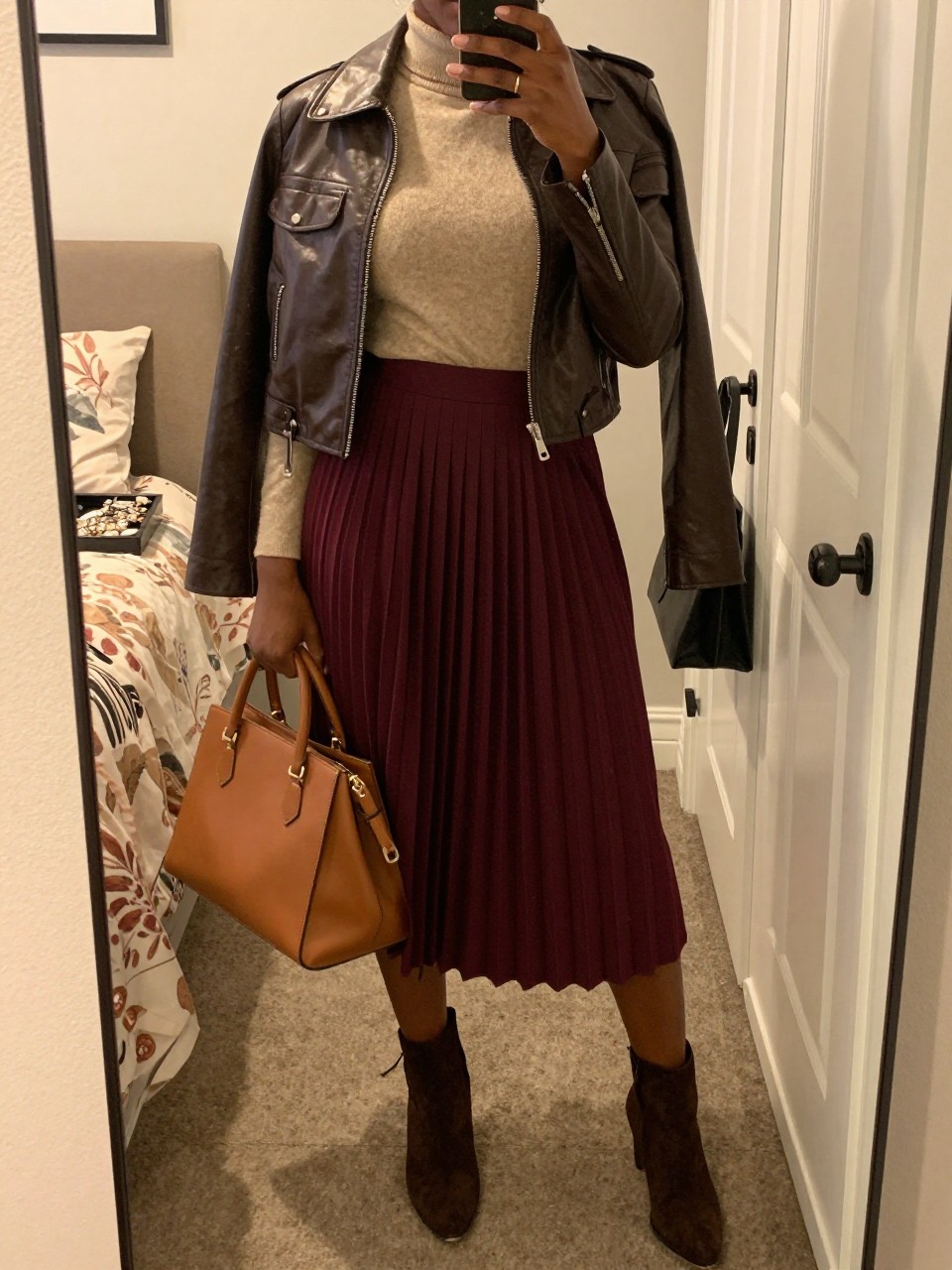 Mirror selfie of a Black woman wearing a burgundy pleated midi skirt, a fitted oatmeal mock-neck sweater, a cropped chocolate-brown leather jacket, dark brown suede ankle boots, and a tan top-handle bag, clearly showing the exact clothing items, colors, fabrics, and accessories listed, holding a phone in front of a feminine-themed bedroom mirror, cropped at the neck so the face does not show, relaxed casual selfie pose, evening golden-hour lighting, a jewelry tray on the vanity and a handbag hanging on a door handle, warm and realistic, latest iPhone photo quality.