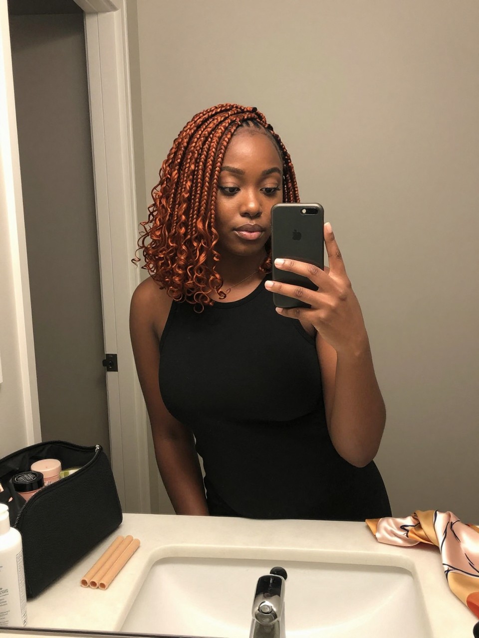 Image prompt: Photo of a Black woman wearing a shoulder-length ginger braided lob with curled tips and a soft side part, candid mirror selfie, bathroom vanity mirror with skincare products and a makeup bag nearby, soft indoor lighting, containing perm rods on the counter and a silk scarf draped beside the sink, realistic finished styling moment, latest iPhone photo quality.