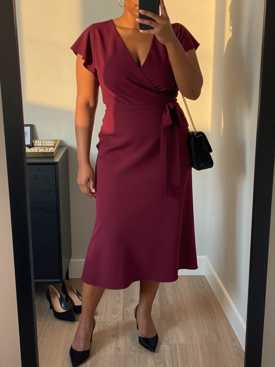 Mirror selfie of a black plus size woman wearing a burgundy crepe wrap midi dress with short flutter sleeves, a modest V-neckline, and a matching waist tie, black pointed slingback heels with low block heels, a small black velvet shoulder bag, and delicate gold hoop earrings, clearly showing the exact clothing items, burgundy crepe, wrap shape, black footwear, and accessories listed, holding a phone in front of a mirror, cropped at the neck so the face does not show, relaxed confident selfie pose, in a stylish city apartment with evening golden-hour lighting, a jewelry tray on the dresser and a pair of heels on the floor nearby, natural unstaged dinner-date appearance, latest iPhone photo quality.