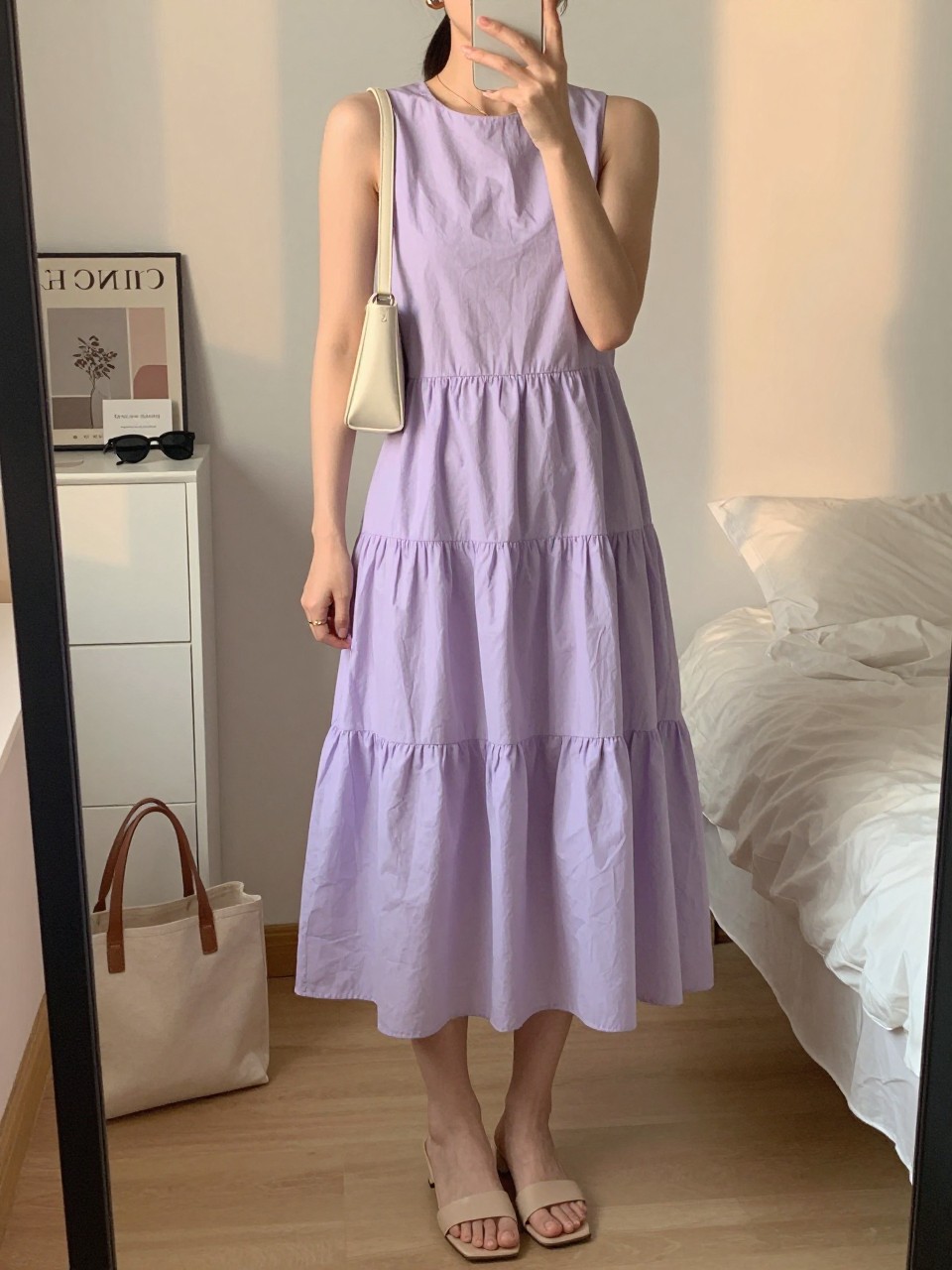 mirror selfie of a woman wearing a soft lilac cotton tiered midi dress with nude leather heeled slides, a cream mini shoulder bag, small gold hoop earrings, and hair in a sleek ponytail, holding a phone in front of a mirror, cropped at NECK so face doesn't show, with a natural casual selfie pose, in a feminine-themed bedroom, evening golden hour, including small lived-in details like a tote bag on the floor, sunglasses on a dresser, or soft bedding in the background, latest iPhone photo quality.