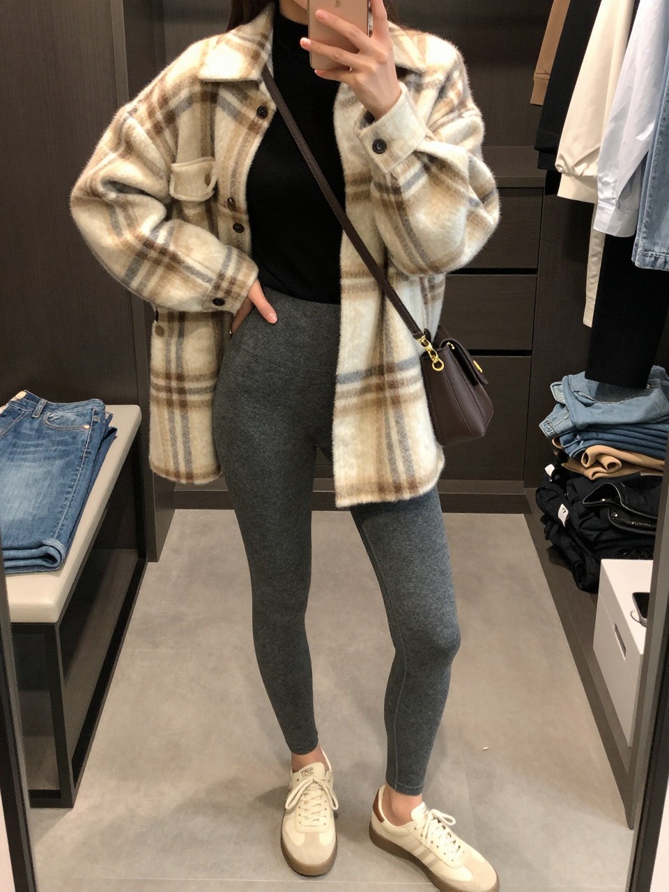 Mirror selfie of a woman wearing charcoal-grey high-waisted leggings, a fitted black mock-neck top, an oversized cream and brown plaid wool-blend shacket, cream leather sneakers, and a dark brown crossbody bag, clearly showing the exact clothing items, colors, fabrics, and accessories listed, holding a phone in front of a walk-in closet mirror, cropped at the neck so the face does not show, relaxed casual selfie pose, soft indoor lighting, open closet with clothes visible and folded denim jeans on a bench, natural and uncluttered, latest iPhone photo quality.