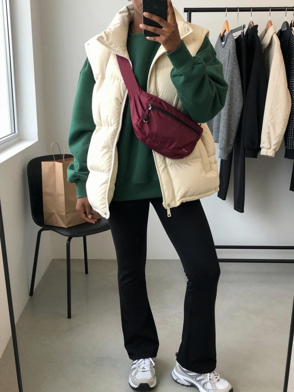 Mirror selfie of a Black woman wearing a cream quilted puffer vest over an oversized forest-green crew-neck sweatshirt, black flared thermal leggings, white and grey trail sneakers, and a burgundy nylon belt bag worn across the chest, clearly showing the exact clothing items, colors, fabrics, and accessories listed, holding a phone in front of a fashion studio mirror corner, cropped at the neck so the face does not show, relaxed casual selfie pose, natural window light, shopping bags beside a chair and a clothing rack with garments behind, believable casual setting, latest iPhone photo quality.