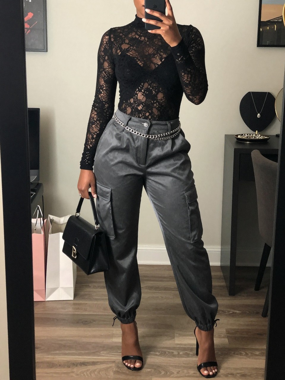 mirror selfie of a Black woman wearing the exact outfit described above, clearly showing a black lace long-sleeve bodysuit, charcoal grey satin cargo pants, black heeled sandals, silver chain belt, and black mini top-handle bag, holding a phone in front of a mirror, cropped at NECK so the face does not show, with a confident casual selfie pose, in a stylish city apartment, soft indoor lighting, including shopping bags beside a chair and a jewelry tray on the vanity, latest iPhone photo quality.