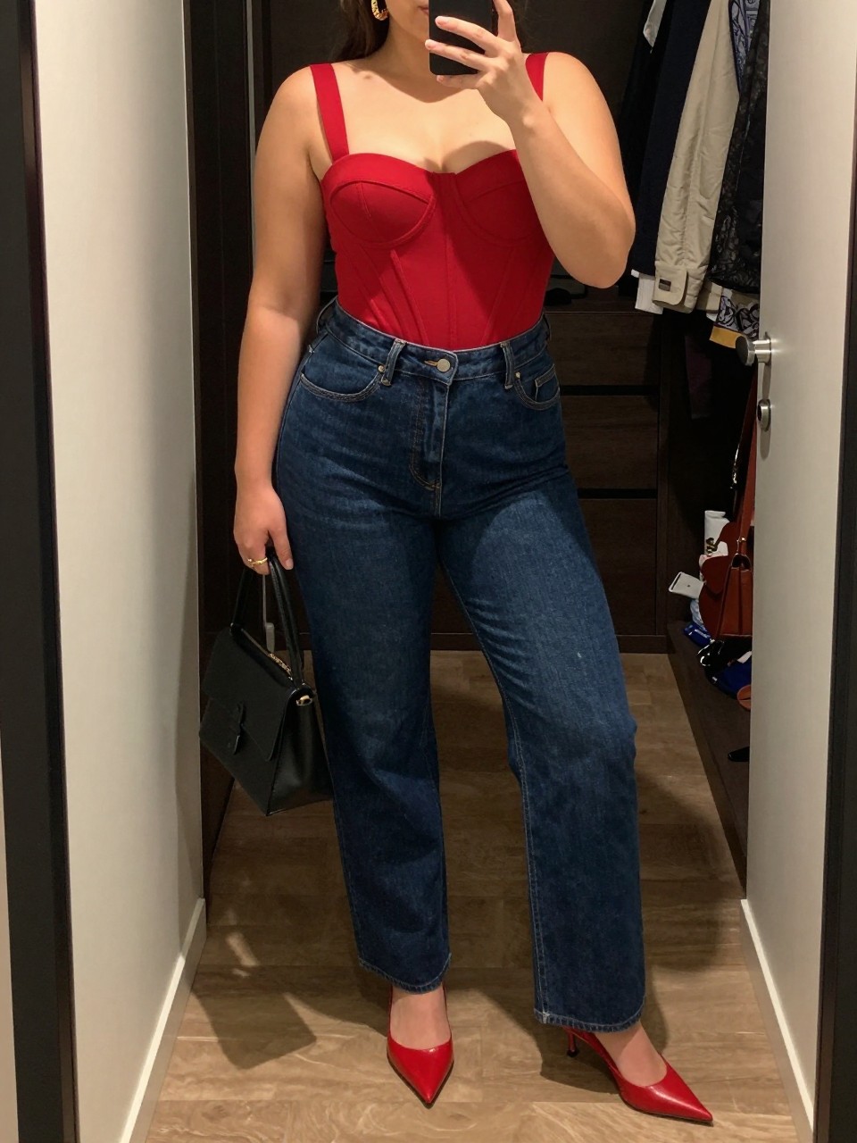 Mirror selfie of a plus size woman wearing dark indigo high-waisted straight-leg jeans, a structured red corset-style top with wide supportive straps, red pointed kitten heels, a small black leather handbag, and chunky gold hoop earrings, clearly showing the exact clothing items, dark denim, red structured top, footwear, and accessories listed, holding a phone in front of a mirror, cropped at the neck so her face does not appear, relaxed bold selfie stance, in a walk-in closet mirror with warm moody lighting, an open closet with clothes visible and a handbag hanging on a door handle, realistic cocktail-date outfit, latest iPhone photo quality.