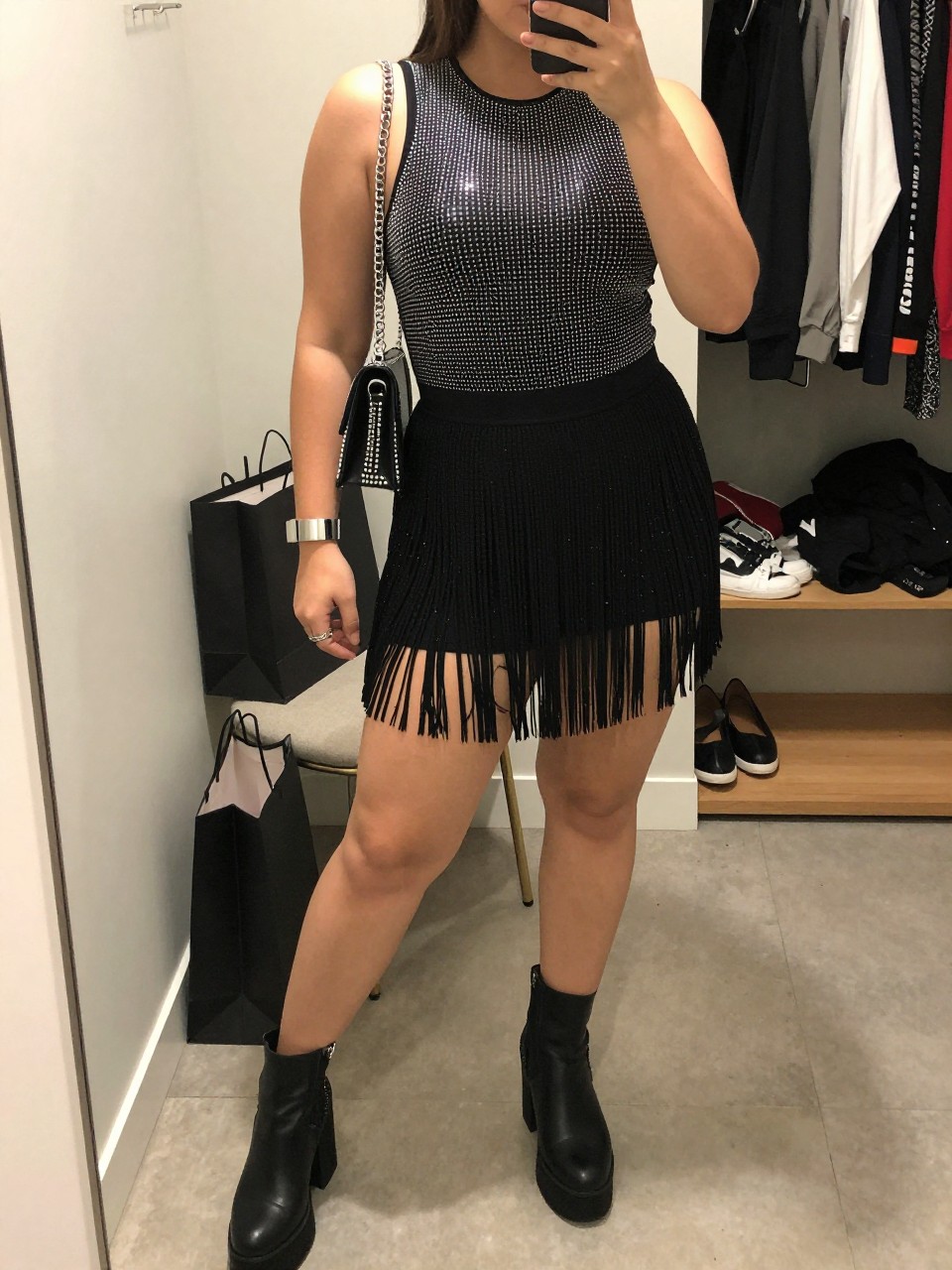 Mirror selfie of a plus size woman wearing a fitted black sleeveless bodysuit covered in fine silver rhinestones, a high-waisted black mini skirt with layered fringe, black platform ankle boots, a small black crystal shoulder bag, and silver ear cuffs, clearly showing the exact clothing pieces, rhinestone details, fringe, colors, footwear, and accessories listed, holding a phone in front of a mirror, cropped at the neck so her face does not appear, relaxed playful selfie stance, in a walk-in closet mirror with soft indoor lighting, shopping bags beside a chair and an open closet with clothes visible, realistic dance-ready club outfit, latest iPhone photo quality.