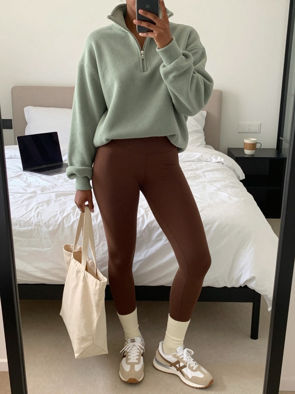Mirror selfie of a Black woman wearing cocoa-brown leggings, an oversized sage-green half-zip fleece sweater, cream crew socks, white and beige retro sneakers, and a beige canvas tote bag, clearly showing the exact clothing items, colors, fabrics, and accessories listed, holding a phone in front of a cozy bedroom mirror setup, cropped at the neck so the face does not show, relaxed casual selfie pose, soft indoor lighting, laptop on the bed and a coffee mug on a bedside table, natural casual atmosphere, latest iPhone photo quality.