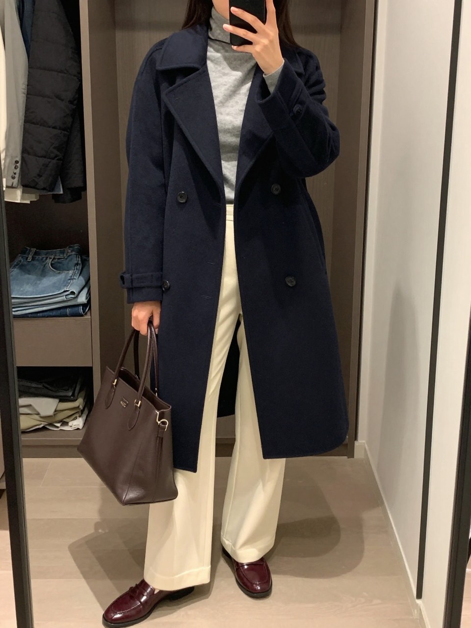 Mirror selfie of a woman wearing a structured navy double-breasted peacoat, fitted heather-grey turtleneck, high-waisted ivory wide-leg trousers, burgundy leather loafers, and a structured dark brown tote bag, clearly showing the exact clothing items, colors, fabrics, and accessories listed, holding a phone in front of a walk-in closet mirror, cropped at the neck so the face does not show, relaxed confident selfie pose, soft indoor lighting, an open closet with clothes visible and folded denim jeans on a bench, natural and polished, latest iPhone photo quality.