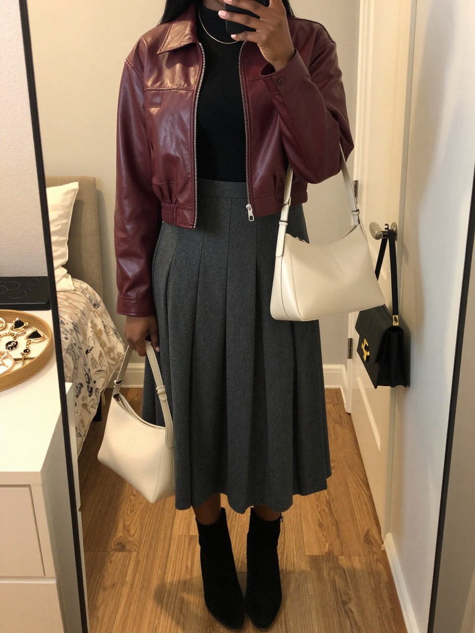 Mirror selfie of a Black woman wearing a cropped burgundy leather jacket, fitted black long-sleeve top, charcoal-grey pleated midi skirt, black suede ankle boots, and a structured cream shoulder bag, clearly showing the exact clothing items, colors, fabrics, and accessories listed, holding a phone in front of a feminine-themed bedroom mirror, cropped at the neck so the face does not show, relaxed casual selfie pose, evening golden-hour lighting, a jewelry tray on the vanity and a handbag hanging on a door handle, warm and realistic, latest iPhone photo quality.
