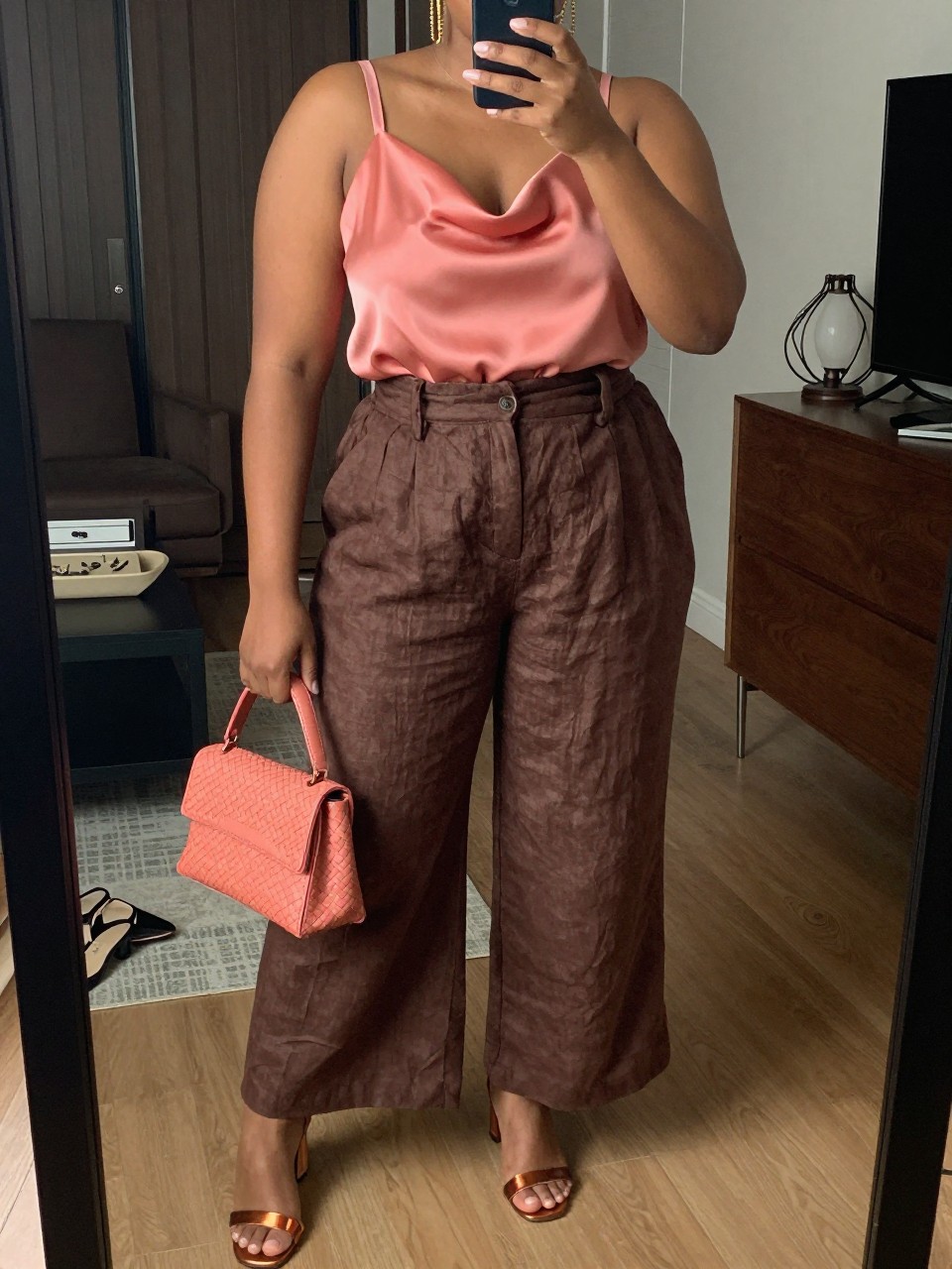 Mirror selfie of a black plus size woman wearing chocolate brown high-waisted palazzo linen pants, a coral pink satin camisole with a softly draped neckline and wide supportive straps, metallic bronze block-heel sandals, a coral woven mini bag, and gold chain earrings, clearly showing the exact clothing items, chocolate linen, coral satin, colors, footwear, and accessories listed, holding a phone in front of a mirror, cropped at the neck so her face is hidden, relaxed elegant selfie pose, in a stylish city apartment with warm moody lighting, a pair of heels on the floor nearby and a jewelry tray on the dresser, realistic summer dinner outfit, latest iPhone photo quality.