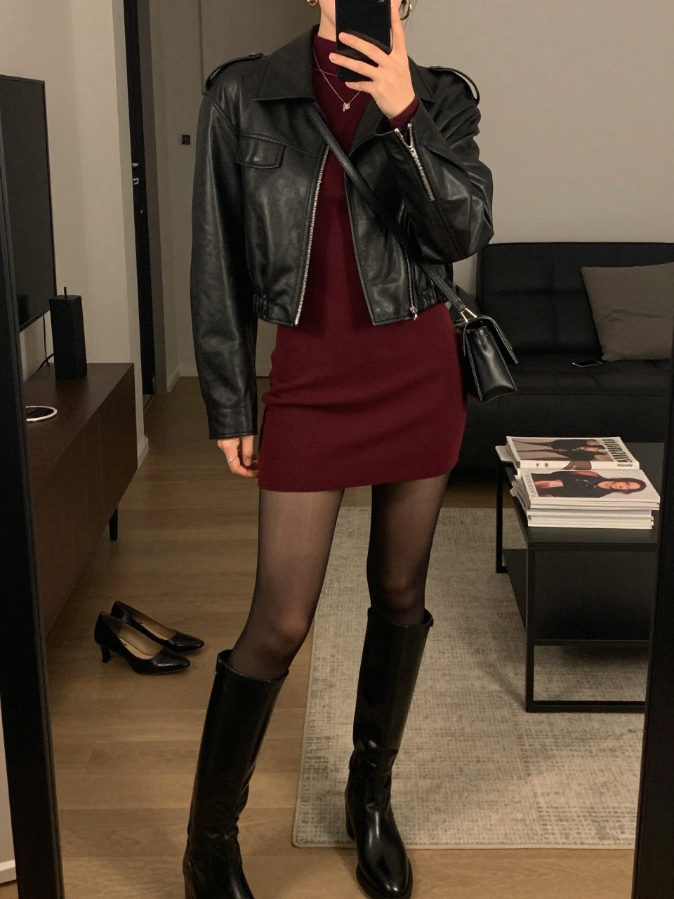 mirror selfie of a woman wearing the exact outfit described above, clearly showing a black cropped leather jacket layered over a burgundy long-sleeve mini dress, black knee-high boots, sheer black tights, a small black crossbody bag, and gold hoop earrings, holding a phone in front of a mirror, cropped at NECK so the face does not show, with a relaxed casual selfie pose, in a stylish city apartment, warm moody lighting, with a pair of heels on the floor nearby and a stack of fashion magazines on a side table, latest iPhone photo quality.
