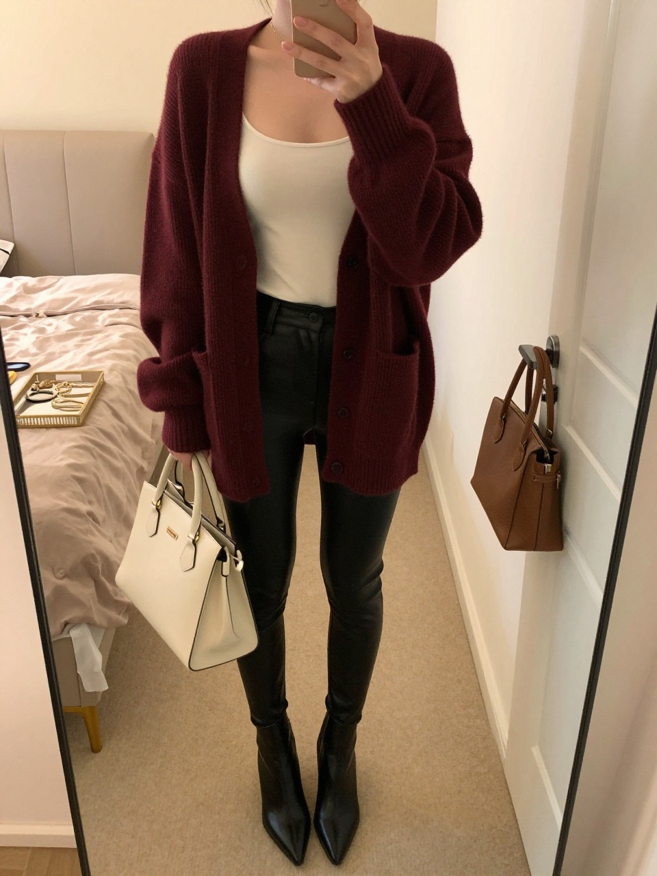 Mirror selfie of a woman wearing black faux-leather leggings, a fitted ivory camisole, an oversized burgundy button-front cardigan, black pointed-toe ankle boots, and a cream structured handbag, clearly showing the exact clothing items, colors, fabrics, and accessories listed, holding a phone in front of a feminine-themed bedroom mirror, cropped at the neck so the face does not show, relaxed casual selfie pose, evening golden-hour light, jewelry tray on the vanity and a handbag hanging on a door handle, warm realistic scene, latest iPhone photo quality.